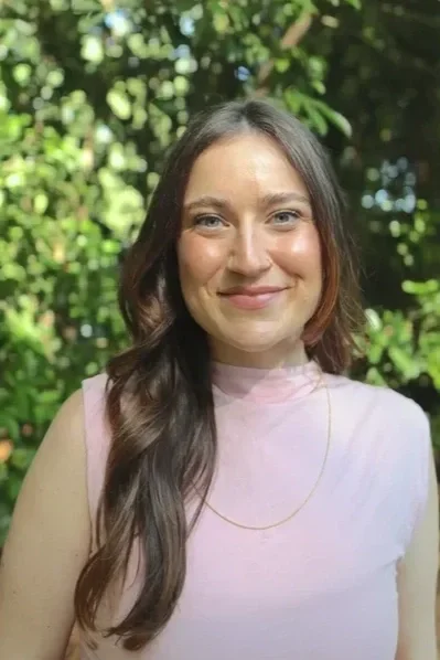 A young woman with long brown hair and fair skin, smiling outdoors with green foliage in the background, wearing a sleeveless pink top and a thin gold chain necklace.
