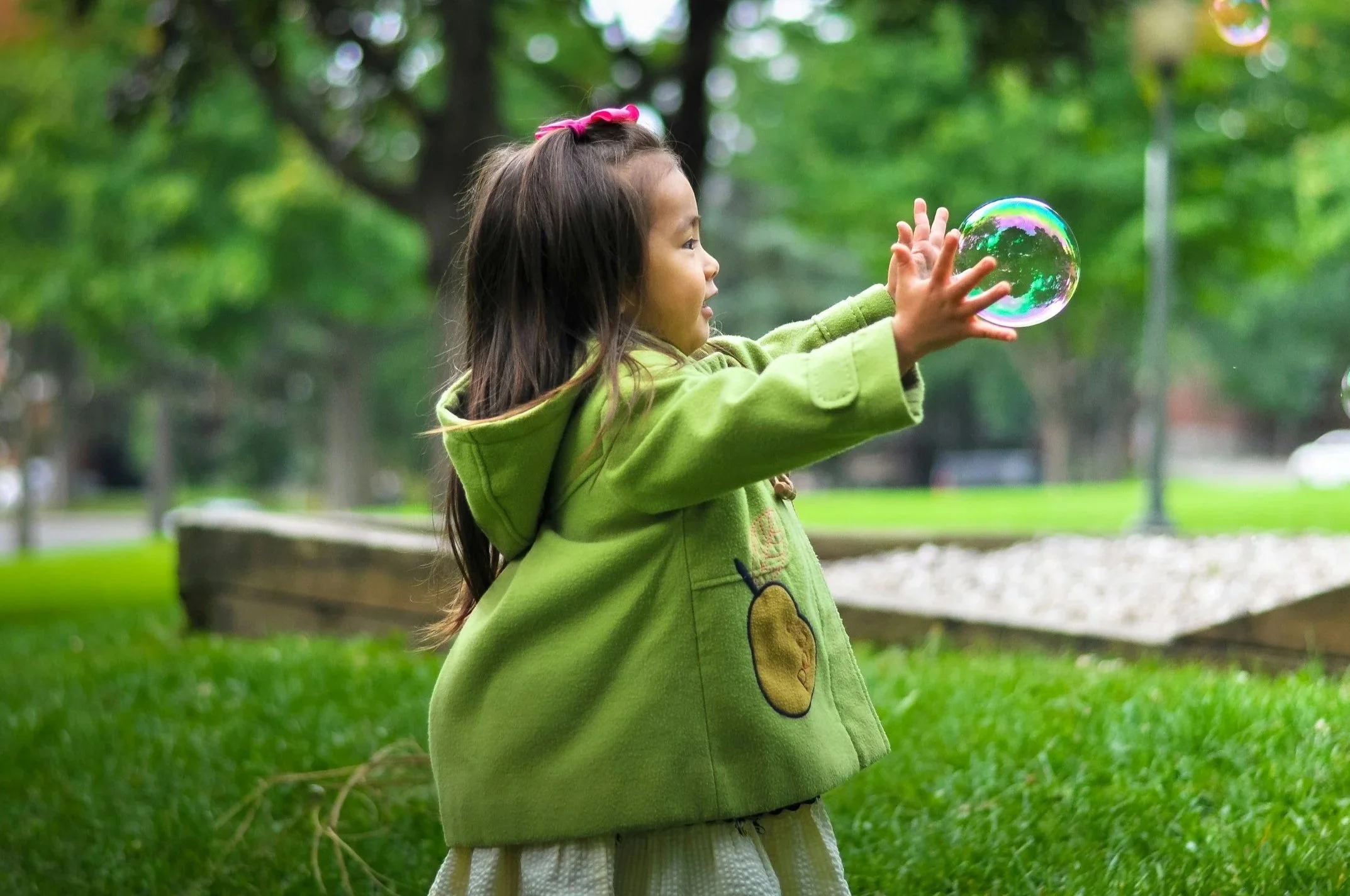 Young girl with long dark hair and a pink bow playing with a soap bubble in a park with green trees and grass.