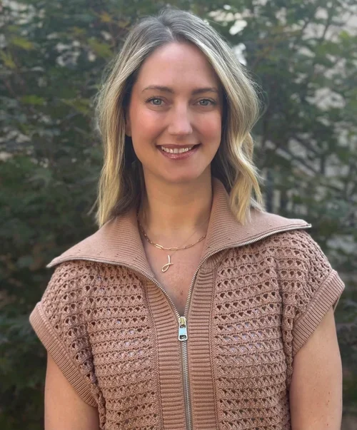 A woman with blonde hair is smiling outdoors, wearing a beige crochet zip-up sweater and a gold necklace with a small pendant, with trees in the background.
