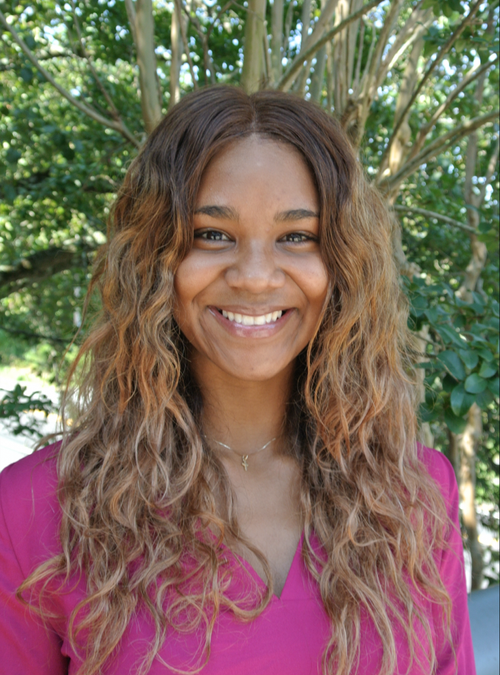 Young woman with curly hair smiling outdoors in front of green trees, wearing a pink top and a delicate necklace.