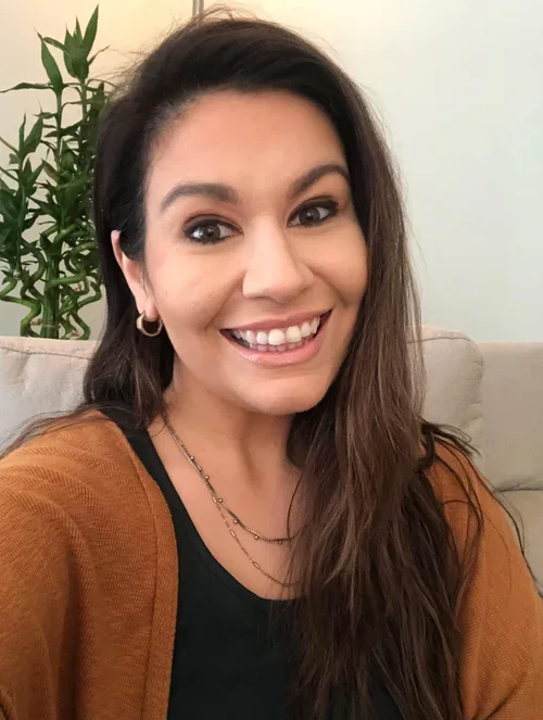 A woman with long brown hair and a smile, wearing a black top and a brown cardigan, sitting on a beige couch with a large green plant in the background.