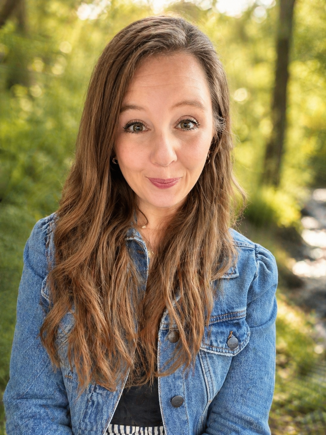 A young woman with long, wavy brown hair and light skin, wearing a denim jacket and smiling, outdoors with green trees and sunlight in the background.