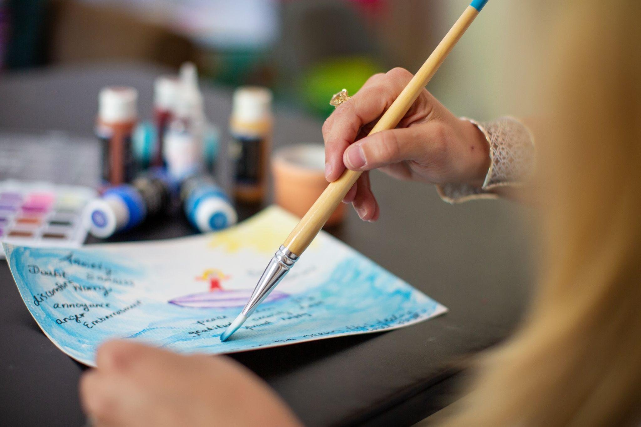 Close-up of a person's hand holding a paintbrush and writing on a colorful piece of paper with handwritten notes, in front of bottles of paint.