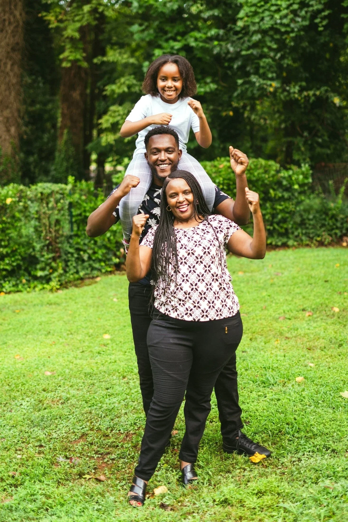 Family of three posing outdoors on a grassy field with trees in the background, with the young girl sitting on her father's shoulders and both adults flexing their arm muscles, smiling cheerfully.