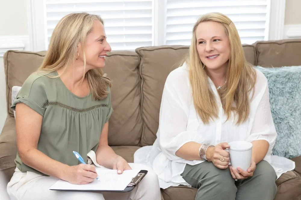 Two women sitting on a beige couch talking, one taking notes and the other holding a mug, in a cozy living room.