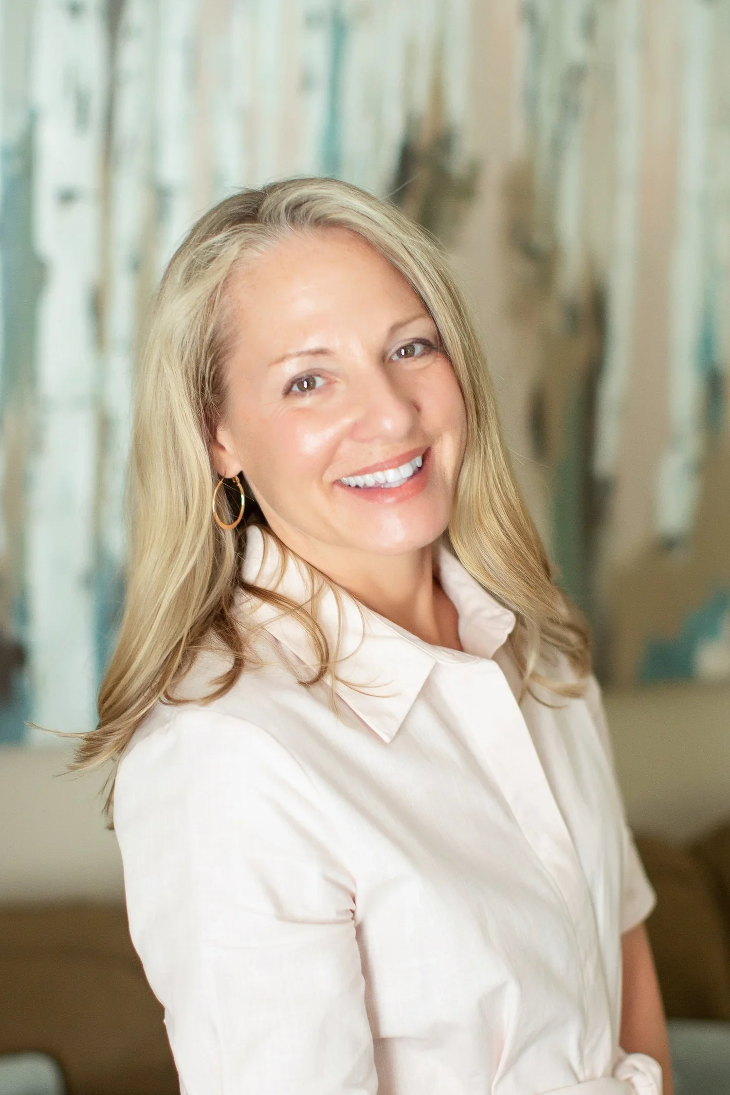 A woman with blonde hair wearing a white blouse, smiling, posed in front of an abstract background.
