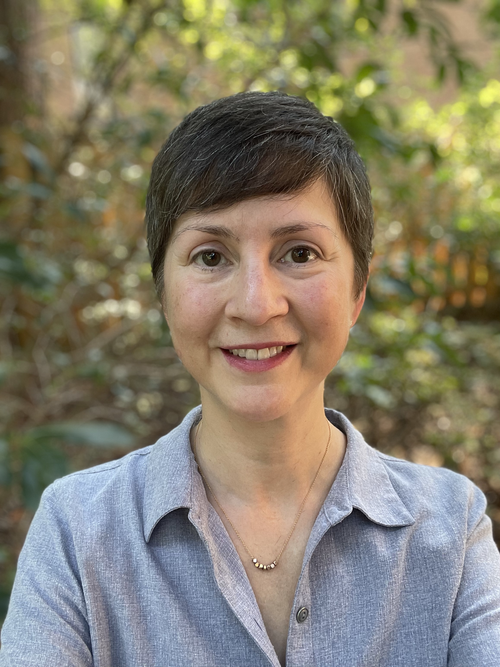A woman with short brown hair wearing a light blue button-up shirt smiling outdoors with blurred trees and foliage in the background.