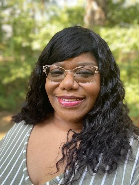 A woman with long curly black hair wearing glasses and a striped shirt, smiling outdoors with trees in the background.