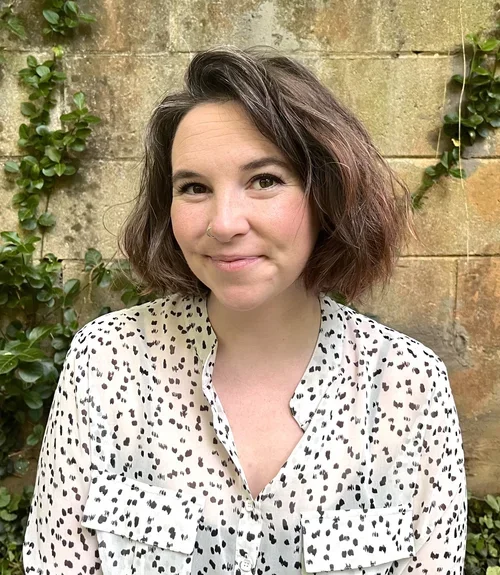 A woman with shoulder-length wavy brown hair and light skin, smiling, in front of a brick wall with green plants.