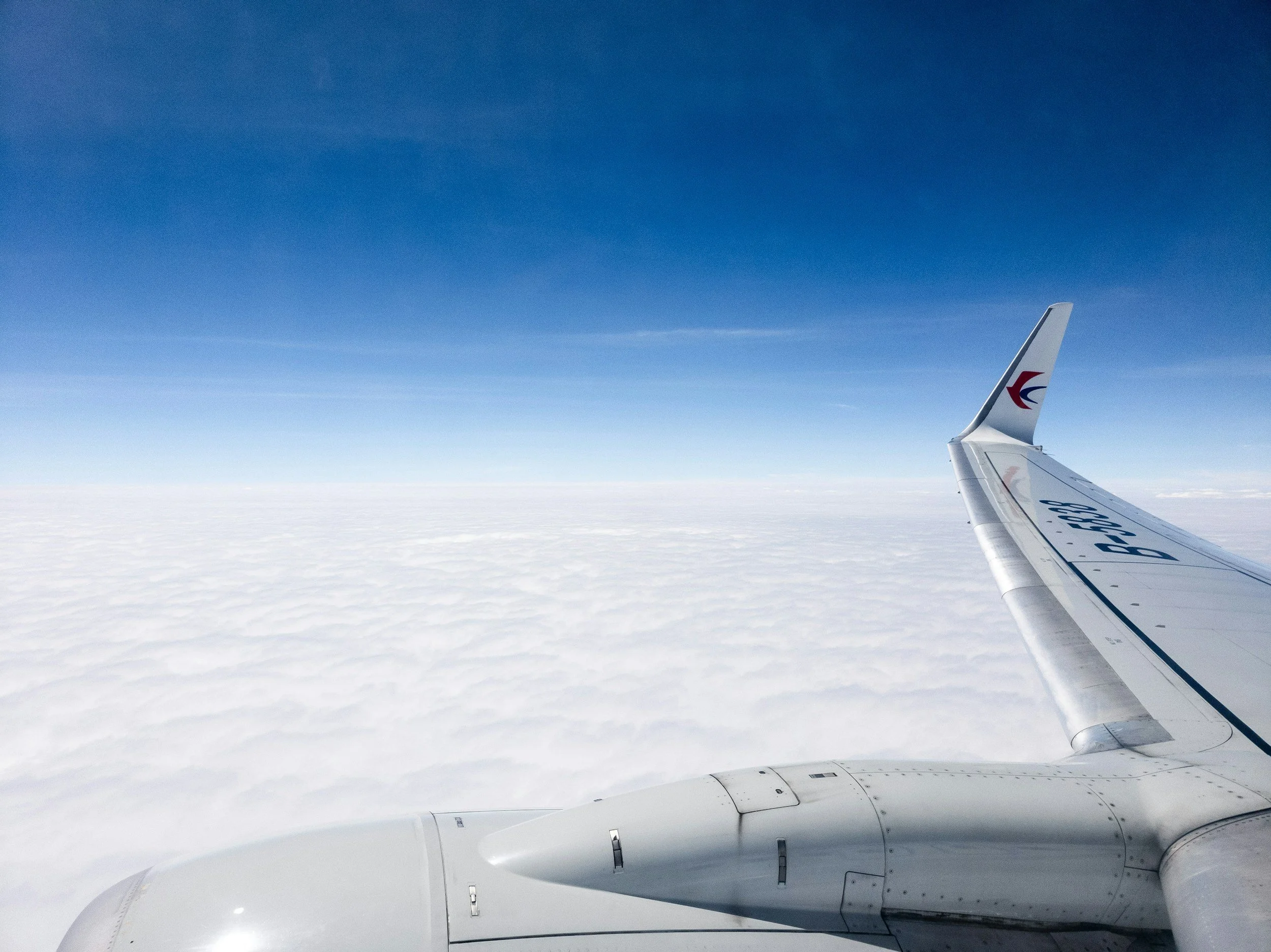 View from an airplane window showing a wing and a sky with clouds below and a clear blue sky above.
