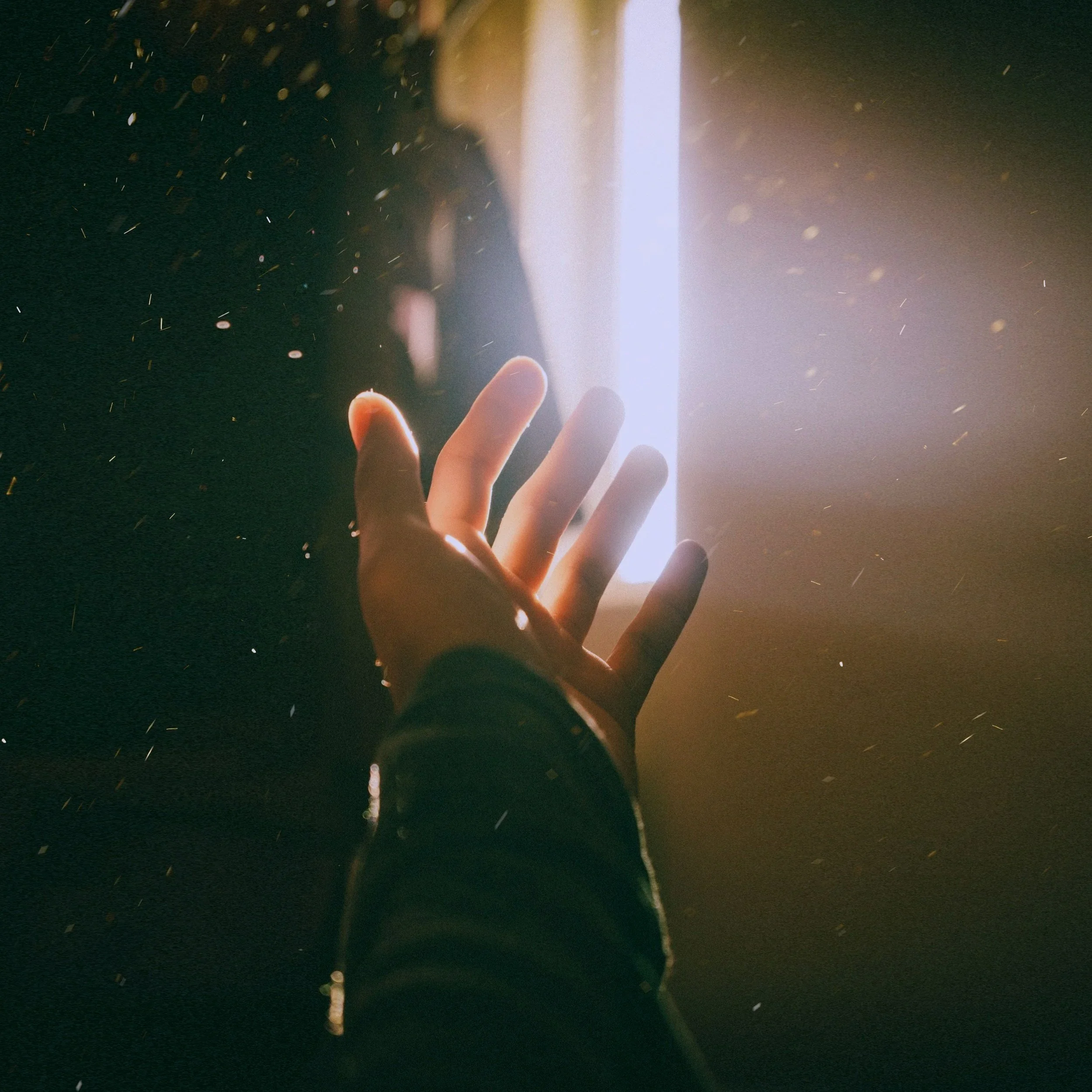 A person's hand with fingers extended, reaching towards a bright light source that is positioned vertically on the wall, illuminating a dark environment with particles or dust floating in the air.