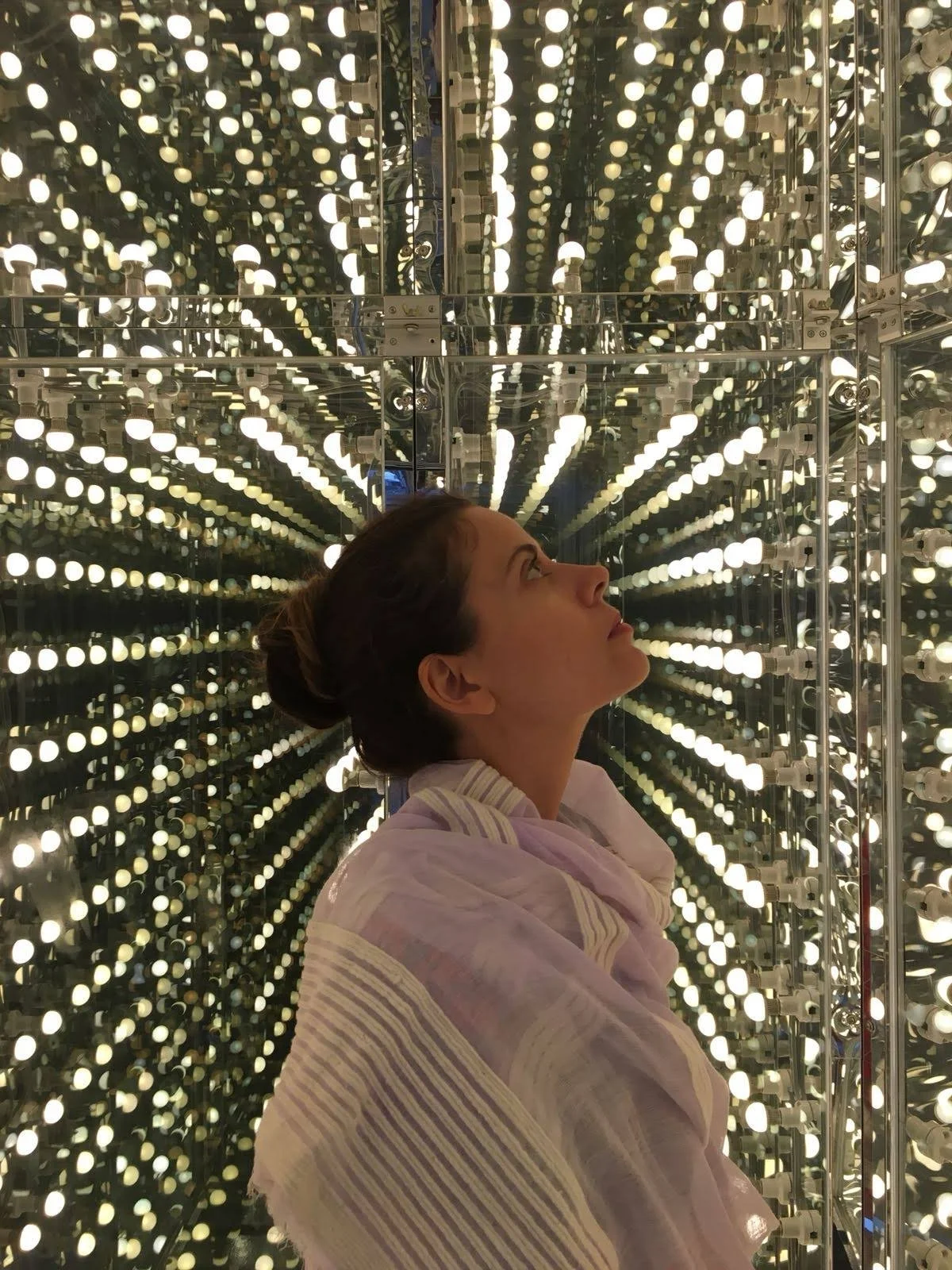 A woman with dark hair tied back in a bun is standing inside a mirrored room surrounded by numerous small, bright lights. She is gazing upward, and her profile is visible. She is wearing a light-colored, striped shawl or blanket.
