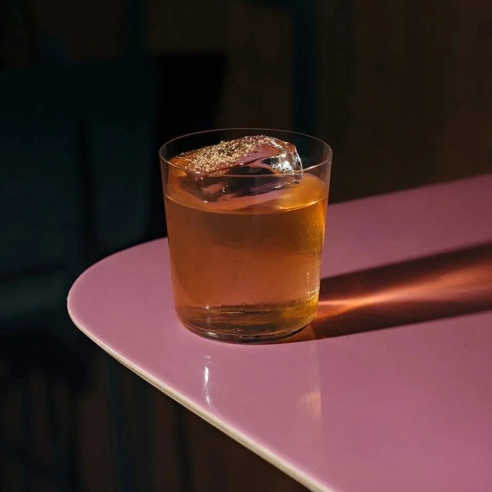 A glass of amber-colored liquid with an ice cube on top, placed on a pink table with a dark background.