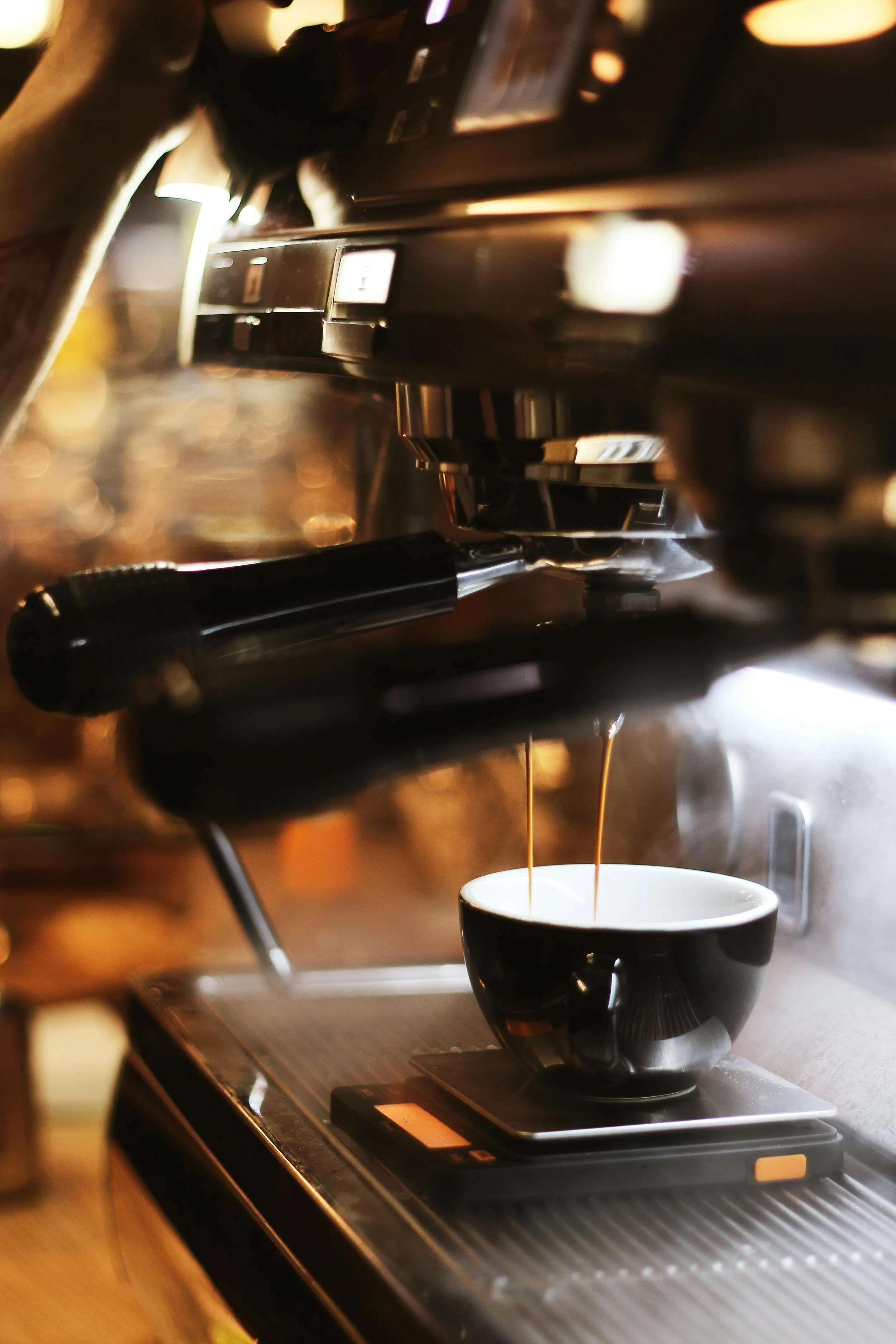Close-up of espresso coffee being brewed in a black and white cup on a scale, with steam rising, under an espresso machine.