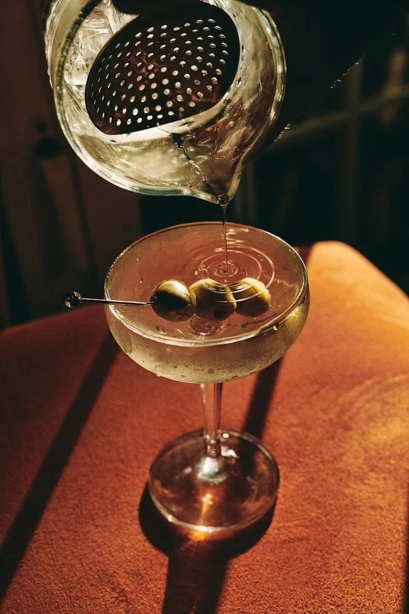 Clear cocktail in a martini glass with green olives, being strained from a shaker topped with a metal strainer.