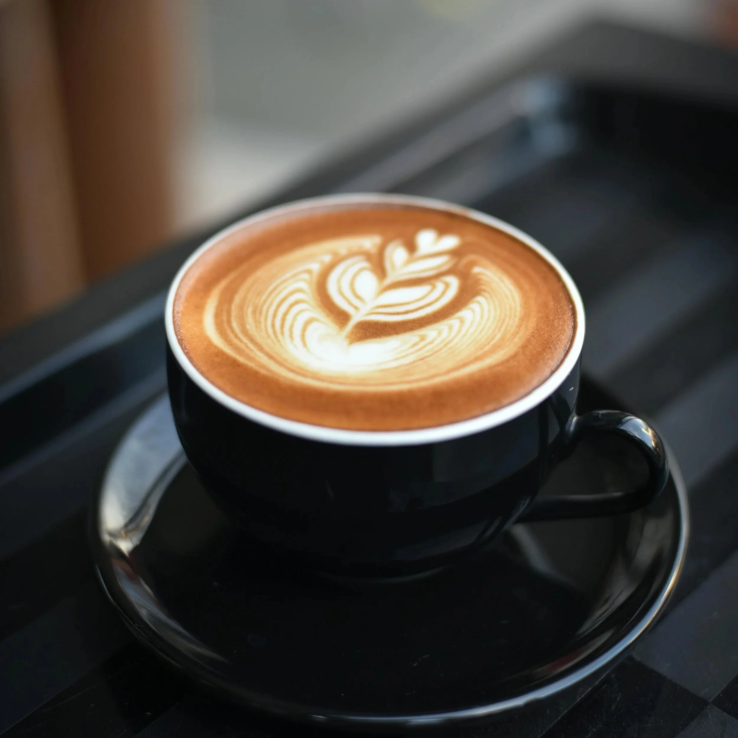 A black cup of coffee with latte art in the shape of a heart and leaves, placed on a matching saucer.