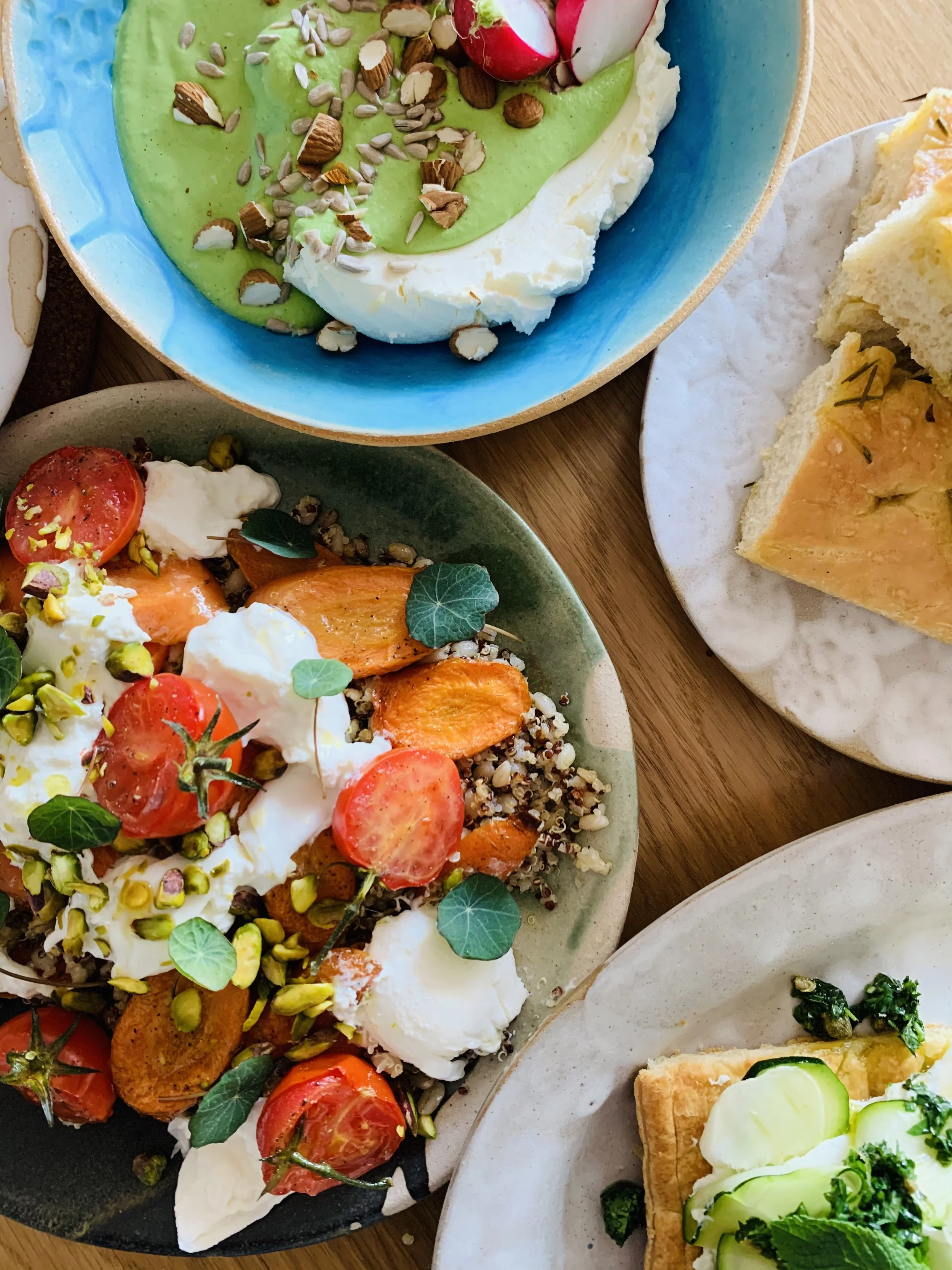 Breakfast dishes including a Greek yogurt bowl with granola, nuts, and fruit; a salad with cherry tomatoes, grilled salmon, boiled eggs, and pistachios; a plate of sliced bread with a spread; and a tofu dish with greens and cucumber.