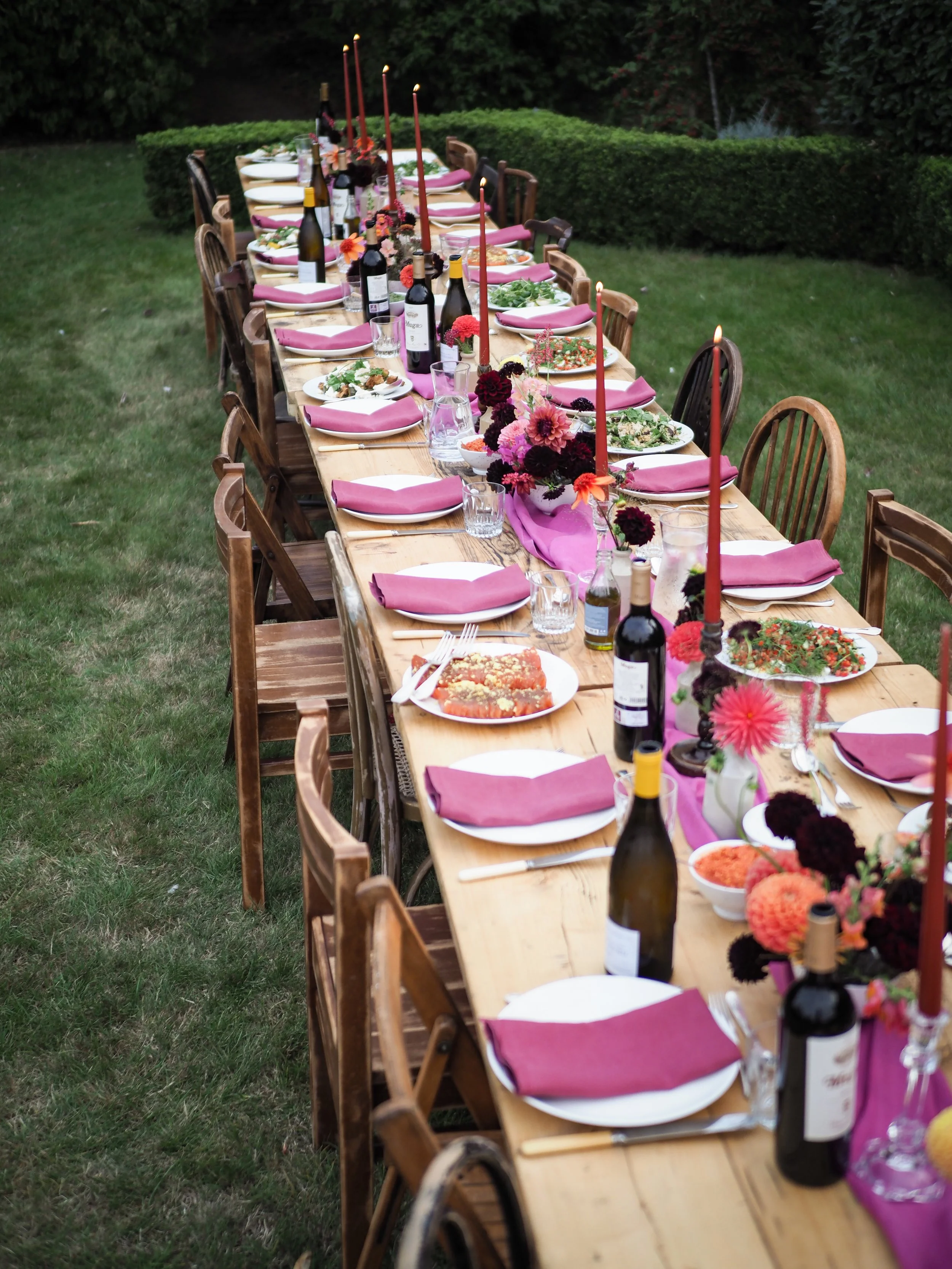 A long outdoor dining table set for a meal, decorated with pink napkins, wine bottles, candles, and floral centerpieces, on a grassy lawn with bushes in the background.