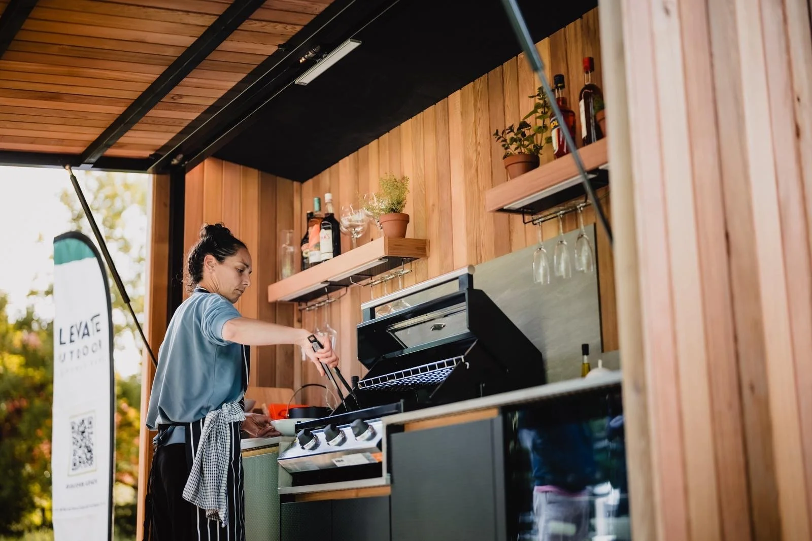 Woman preparing food on an outdoor kitchen with wooden walls, shelves holding liquor bottles, wine glasses, and potted plants.