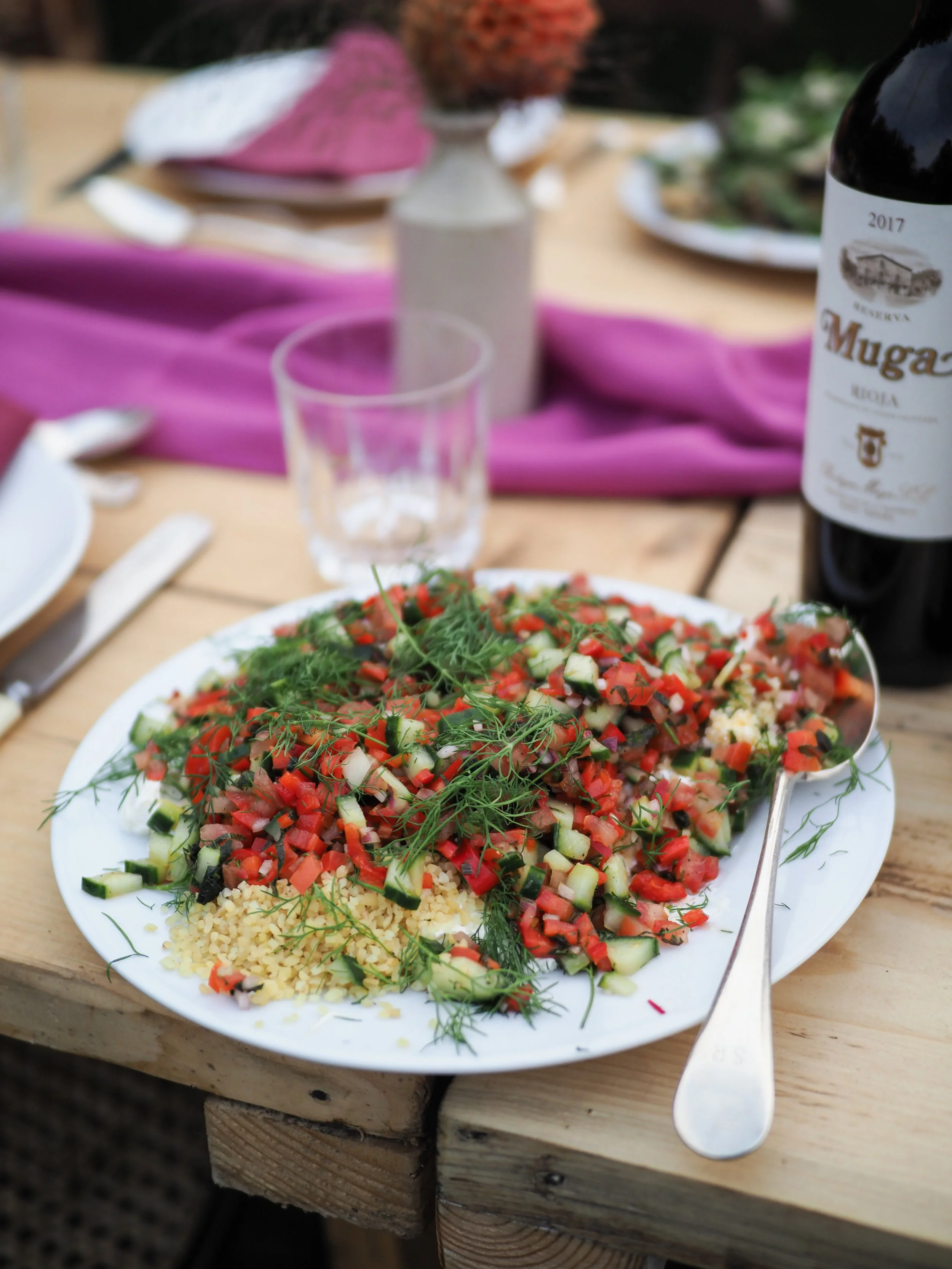 A plate of chopped vegetables with herbs, likely a salad, on a wooden table with a fork. In the background, there is a bottle of red wine, a glass, and a table setting with pink and purple cloths and a vase with flowers.