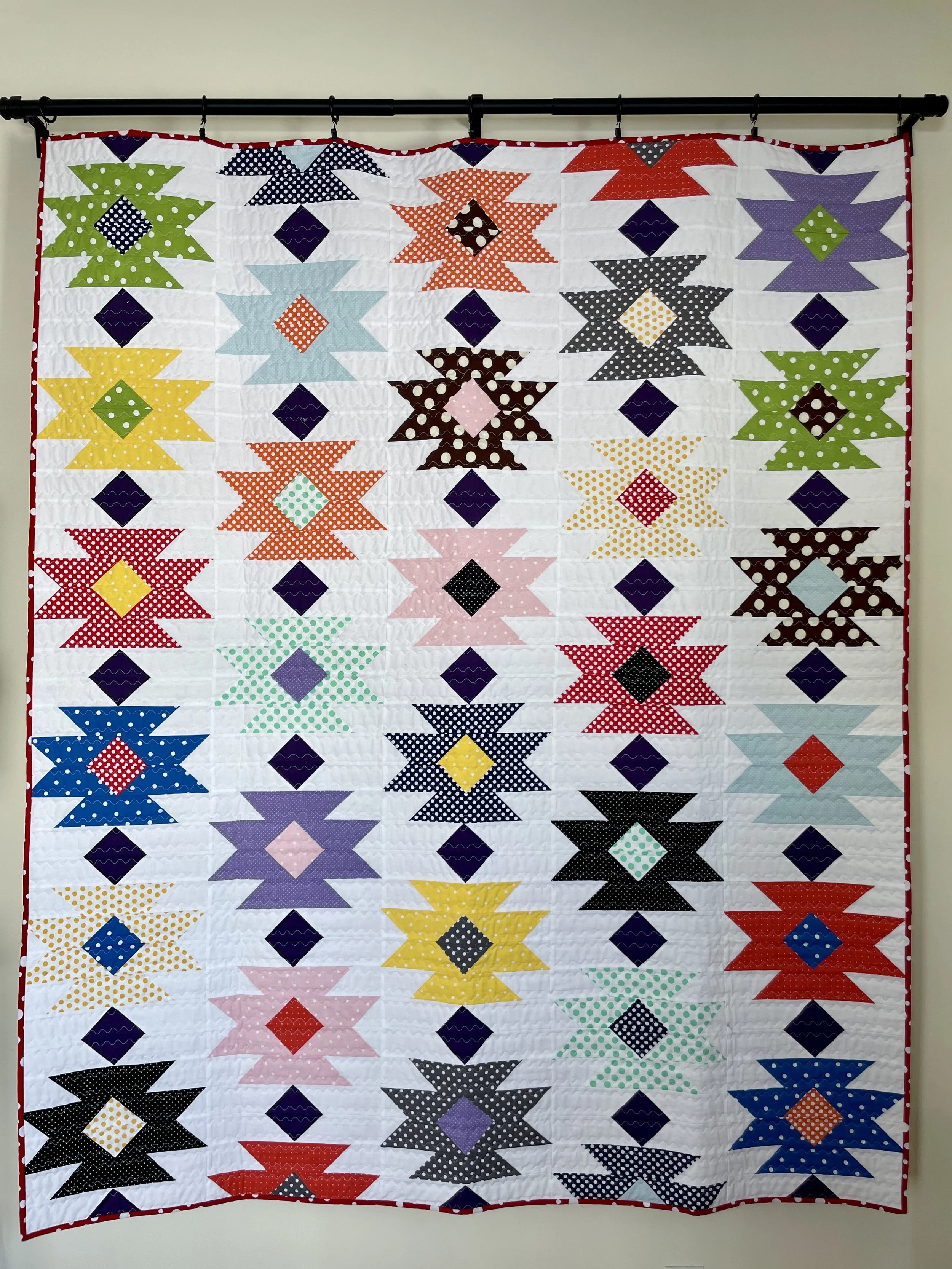 Colorful quilt hanging on a black rod, featuring a pattern of star shapes in various colors and patterns, set against a white background.