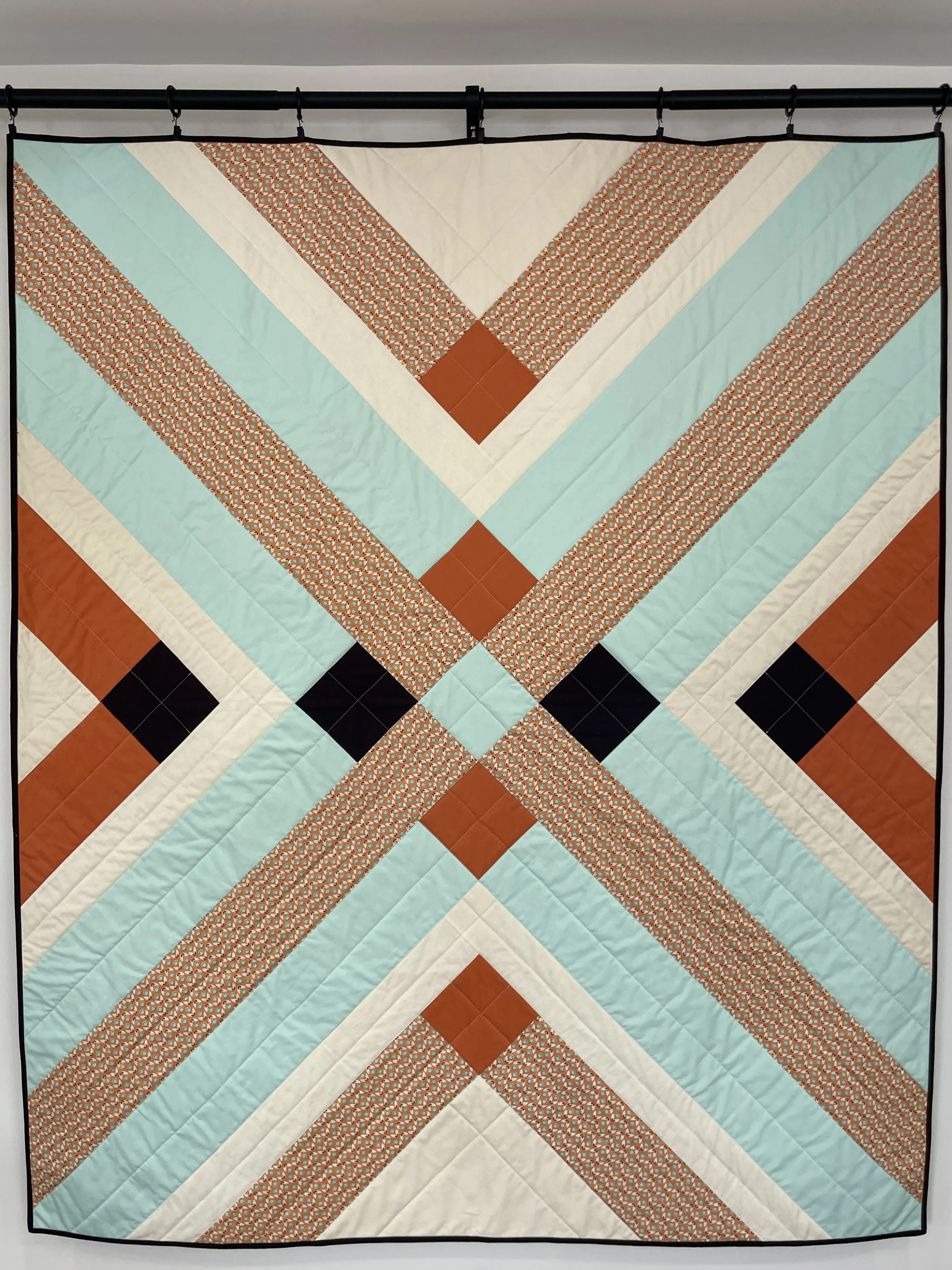 A colorful quilt with geometric patterns in shades of orange, light blue, brown, and white, hanging on a black rod.