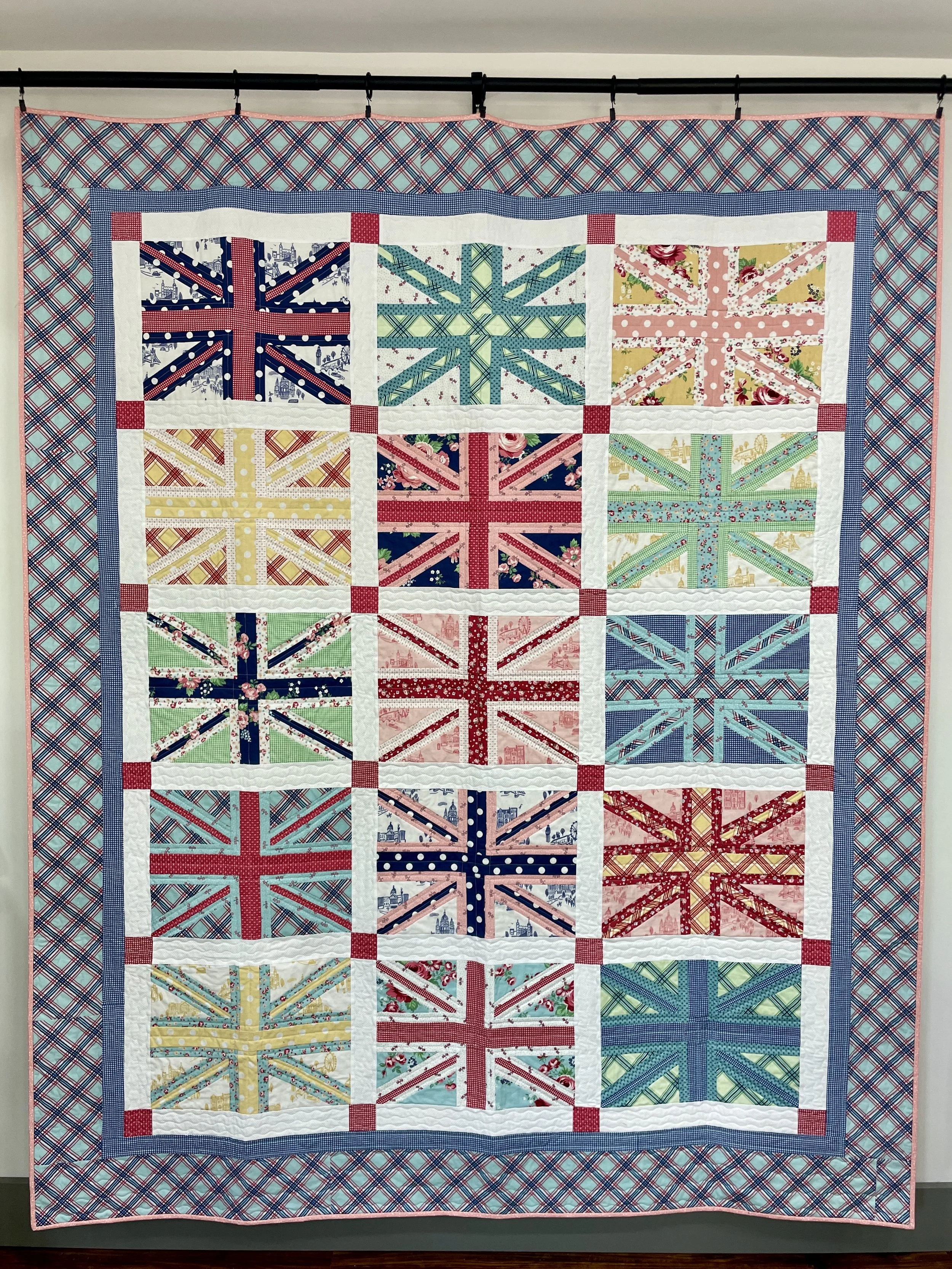 A quilt with a 3x4 grid of blocks, each featuring a Union Jack design in various colorful fabrics, surrounded by a border with a red and blue plaid pattern.