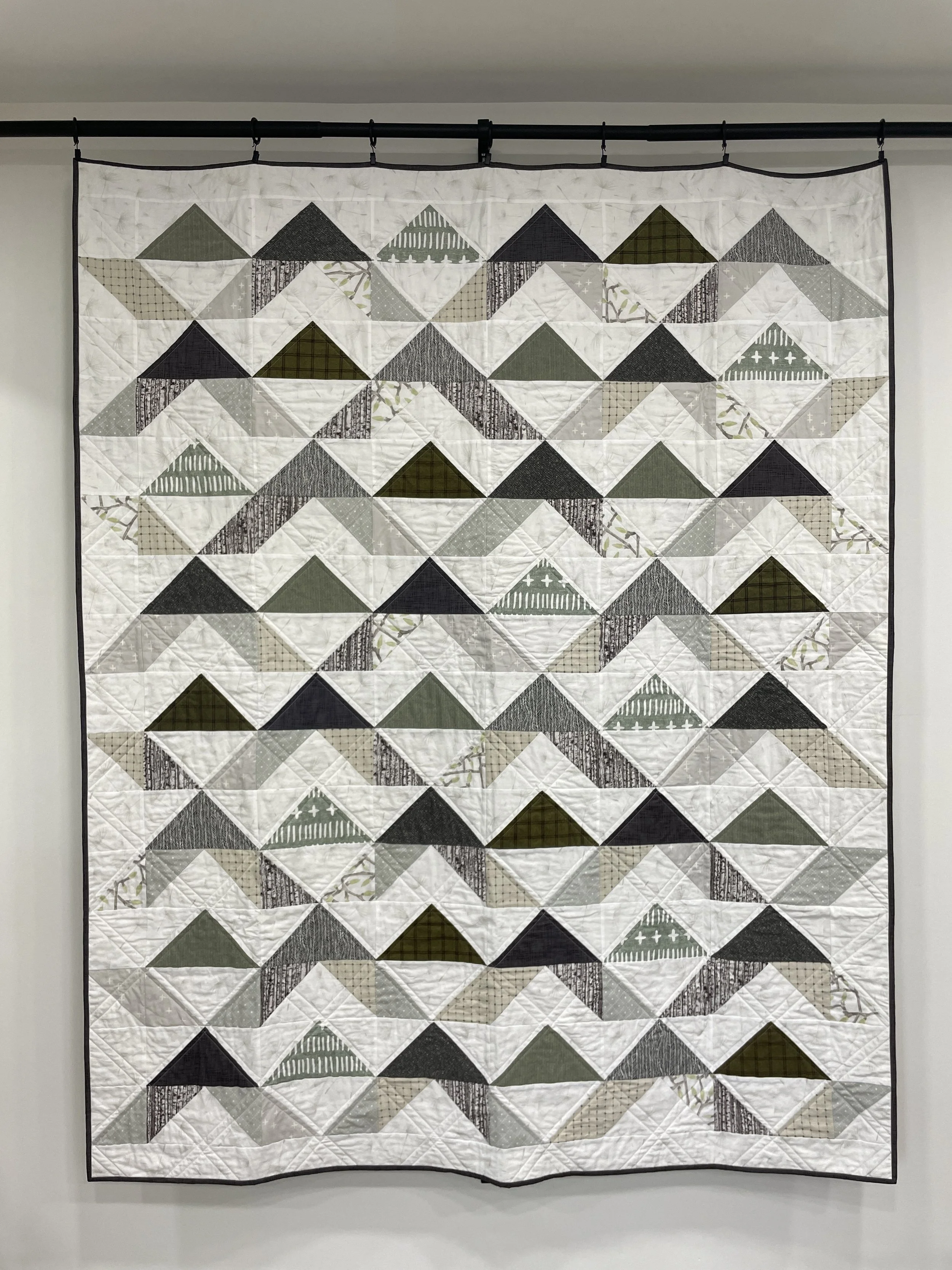 A quilt with a geometric mountain pattern in shades of white, gray, black, and green hung on a wall.