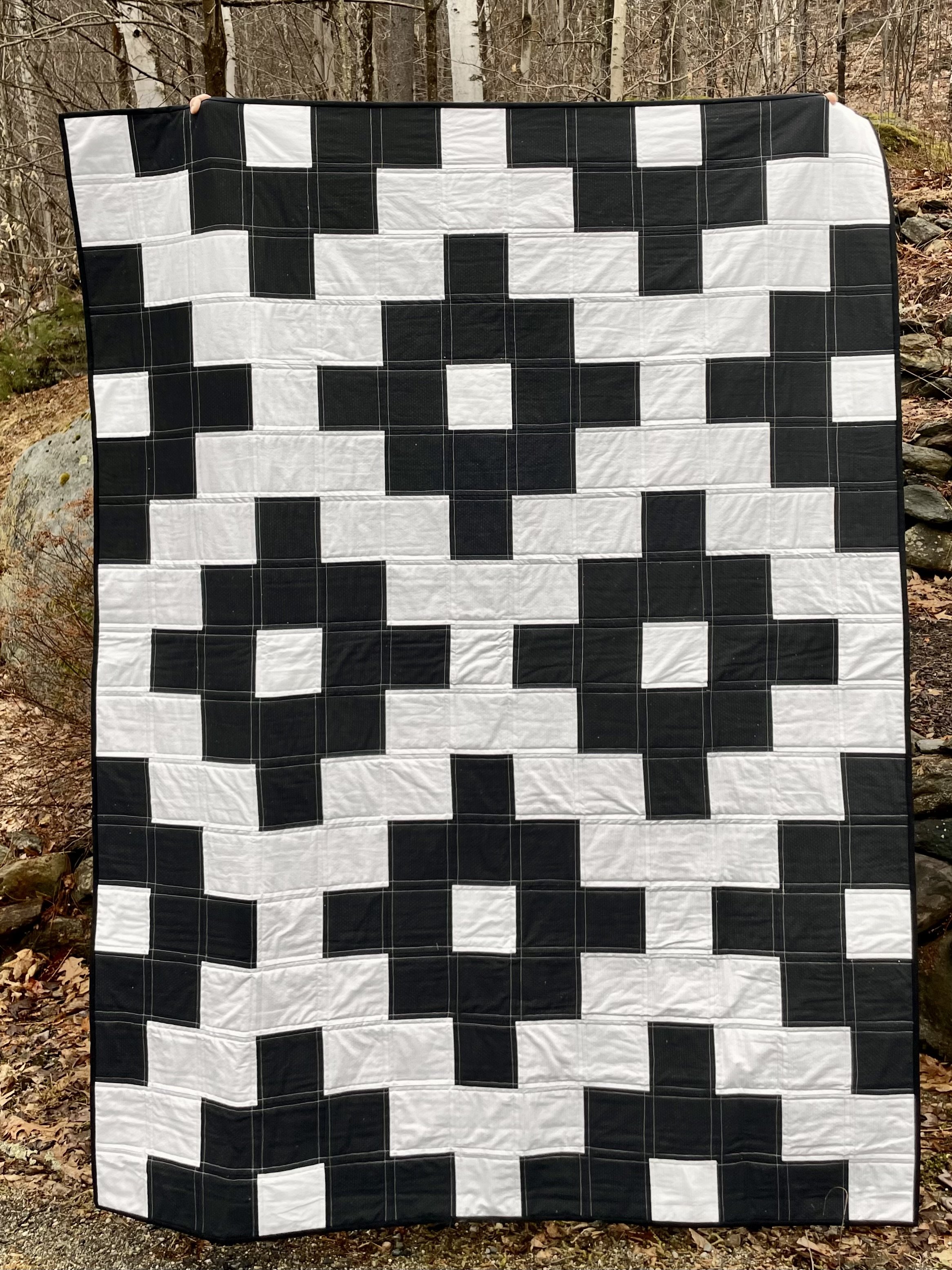A black and white quilt with a pixelated pattern, displayed outdoors with trees and rocks in the background.