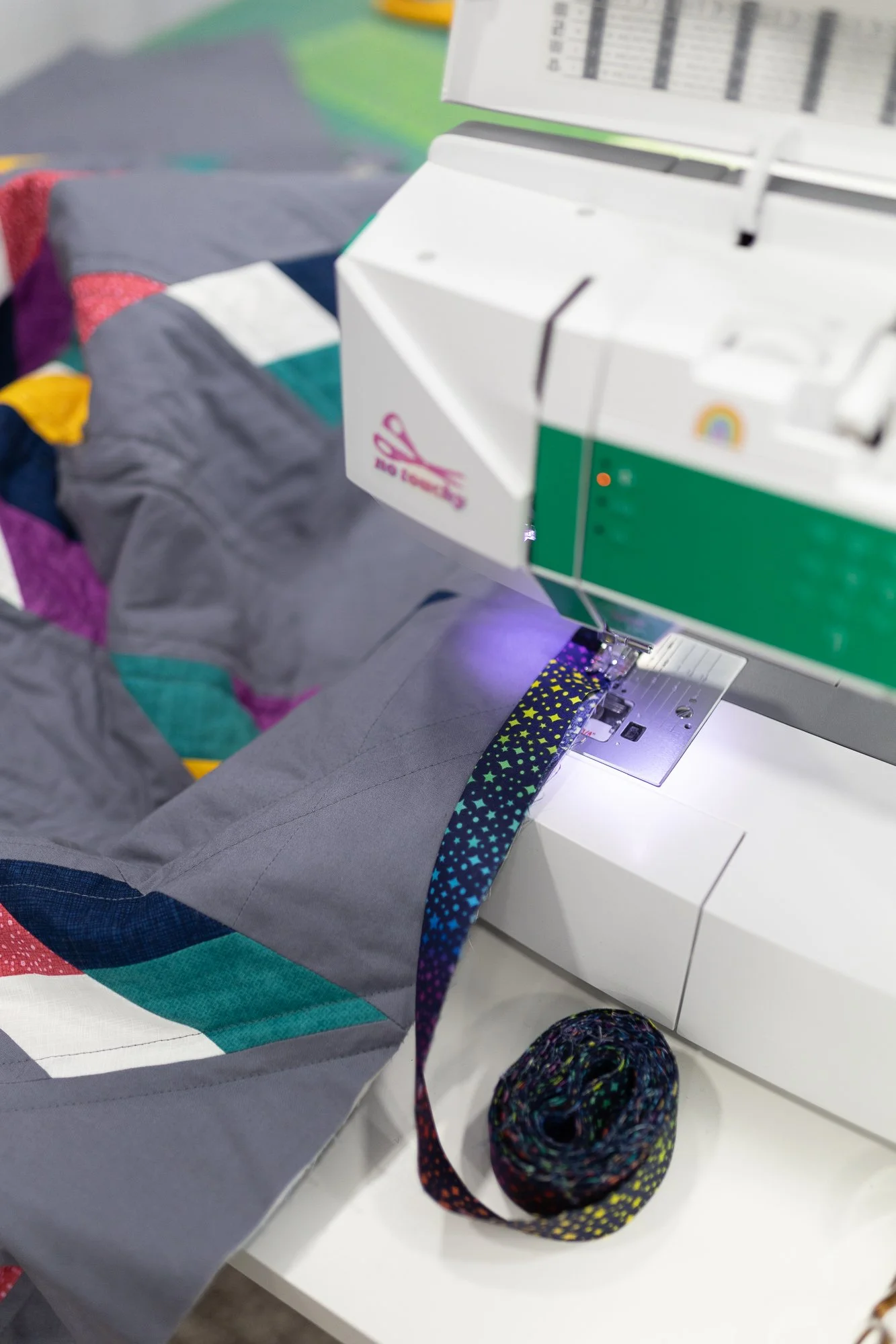 A sewing machine stitching a colorful, patterned fabric with a rainbow logo on the front, beside a spool of multicolored thread, on a white table surrounded by various fabric pieces.