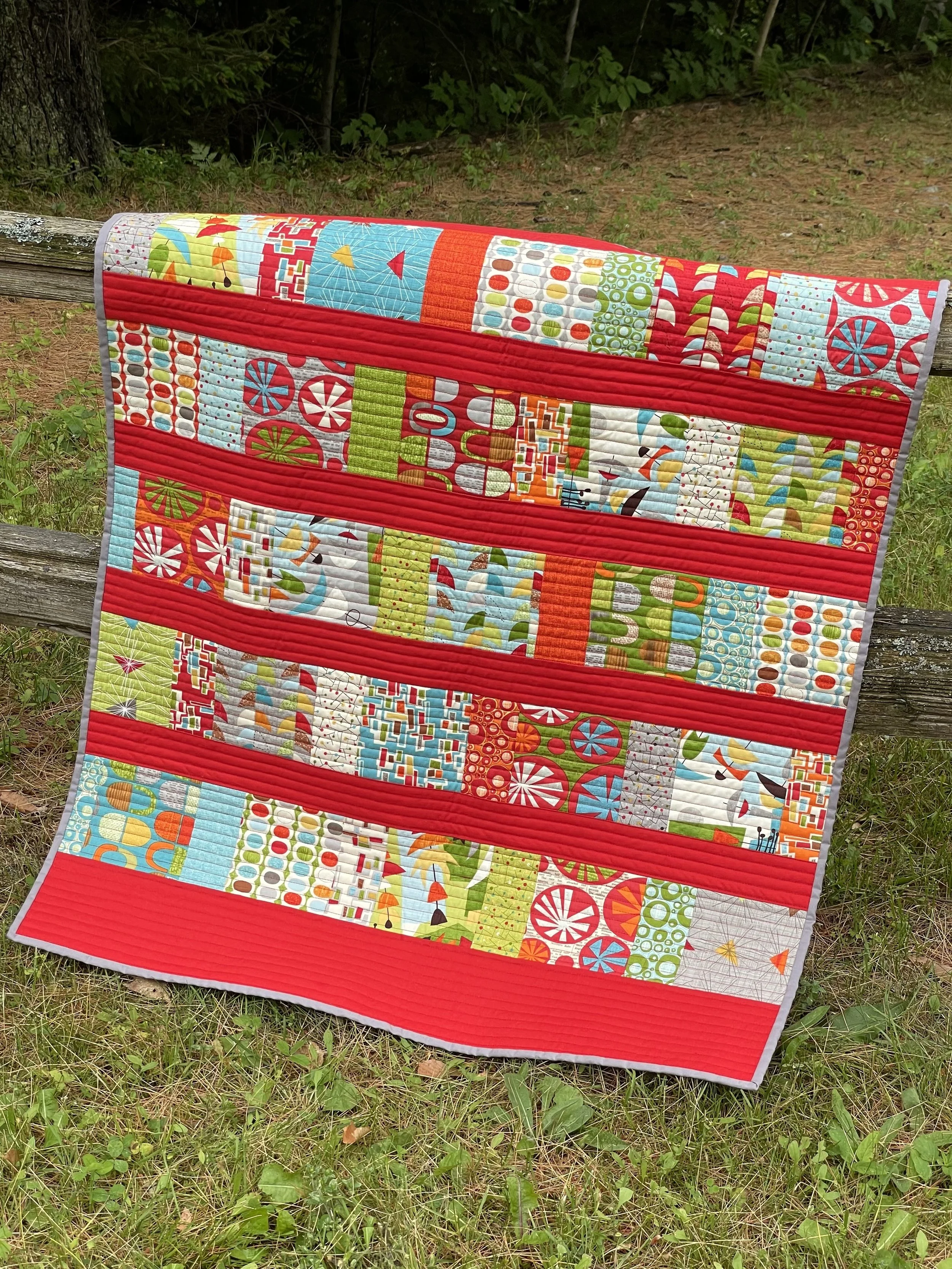 Colorful quilt with horizontal red stripes, hanging on a wooden fence in an outdoor grassy area.