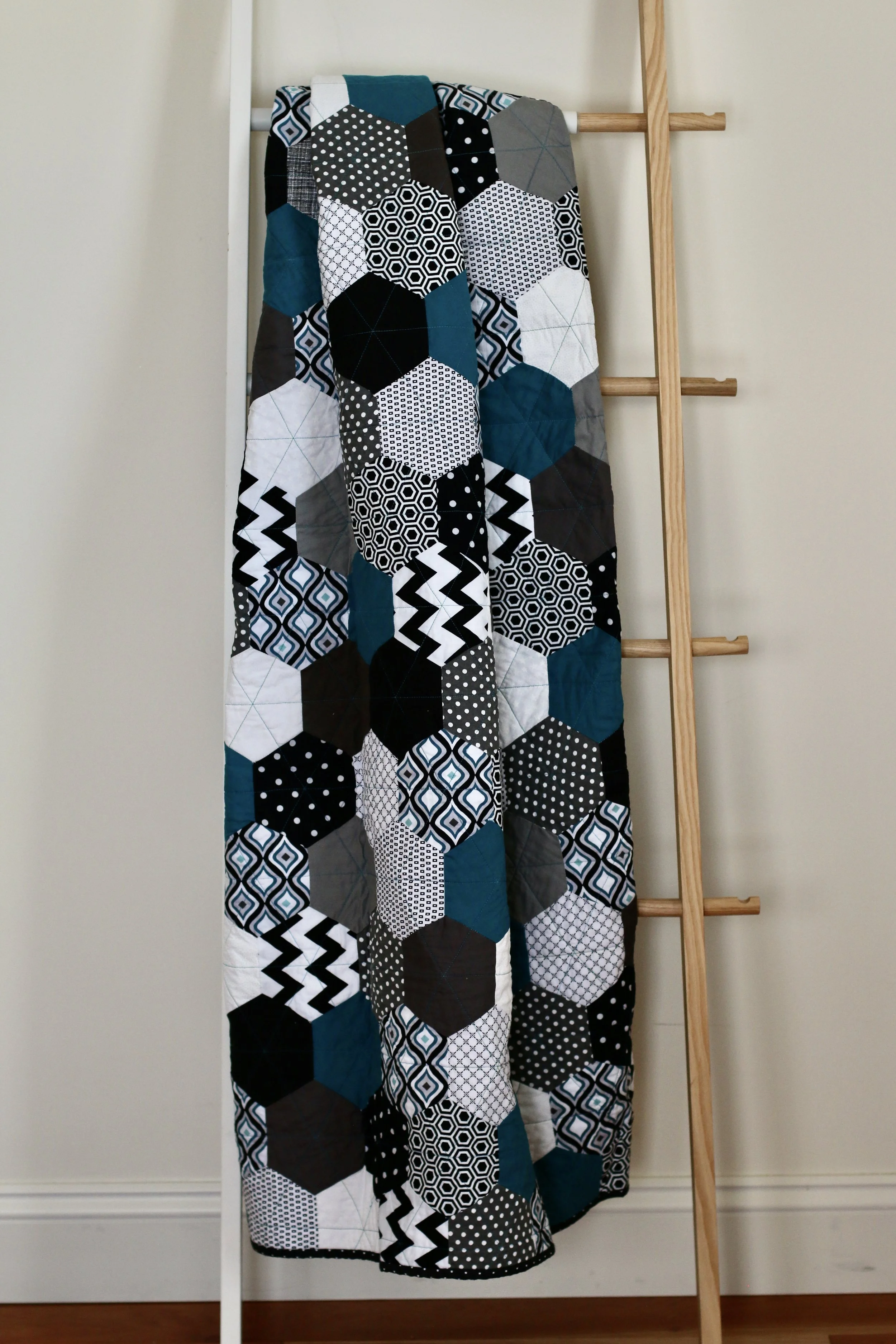 A geometric quilt with hexagon patches in black, white, gray, and teal, hanging on a white ladder against a white wall.