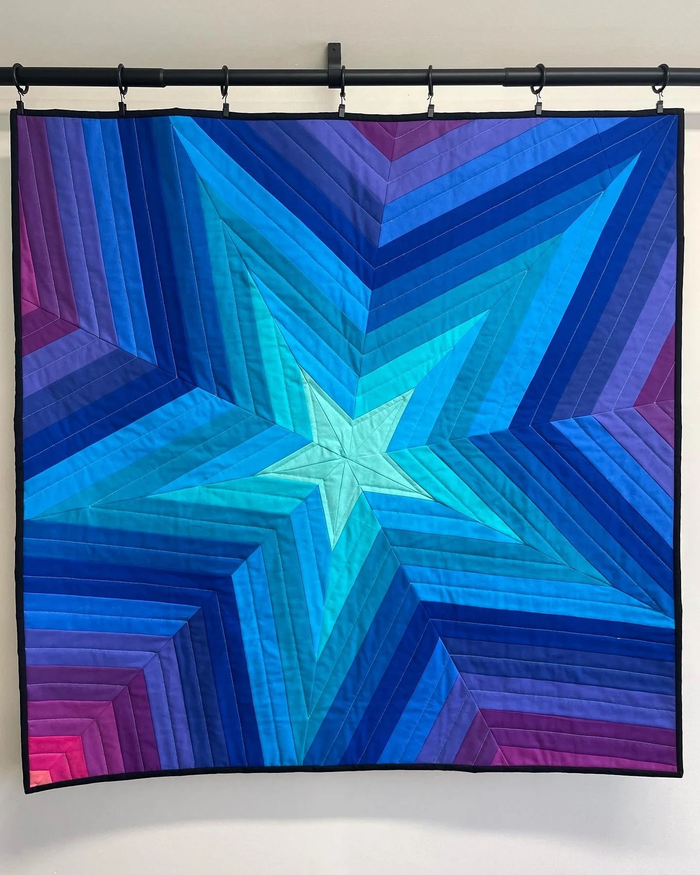 Blue, purple, and pink quilt with a star pattern, hanging on a black rod against a white wall.