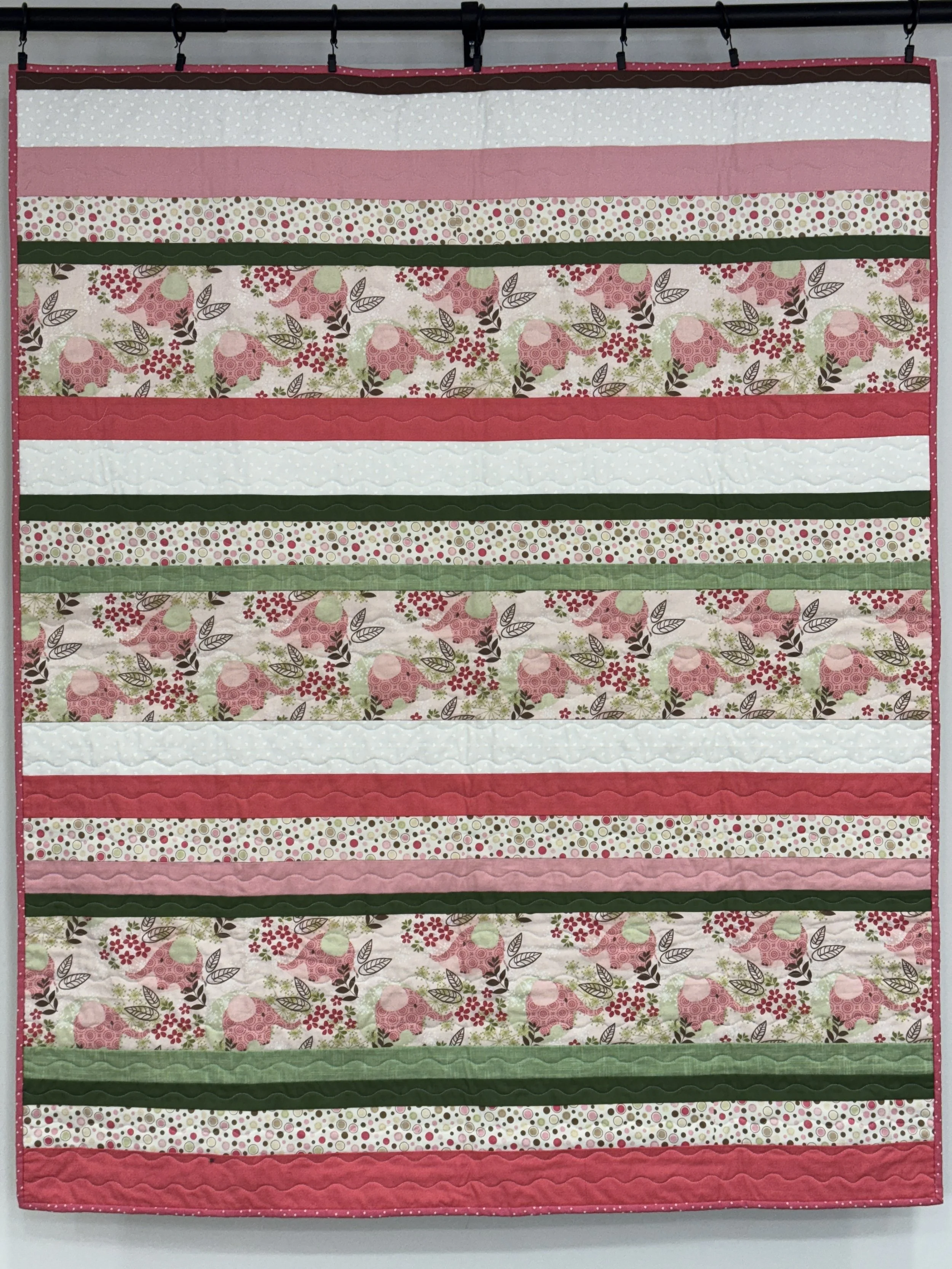 A quilt with horizontal strips in pink, green, white, and patterned fabrics, hanging on a wall.