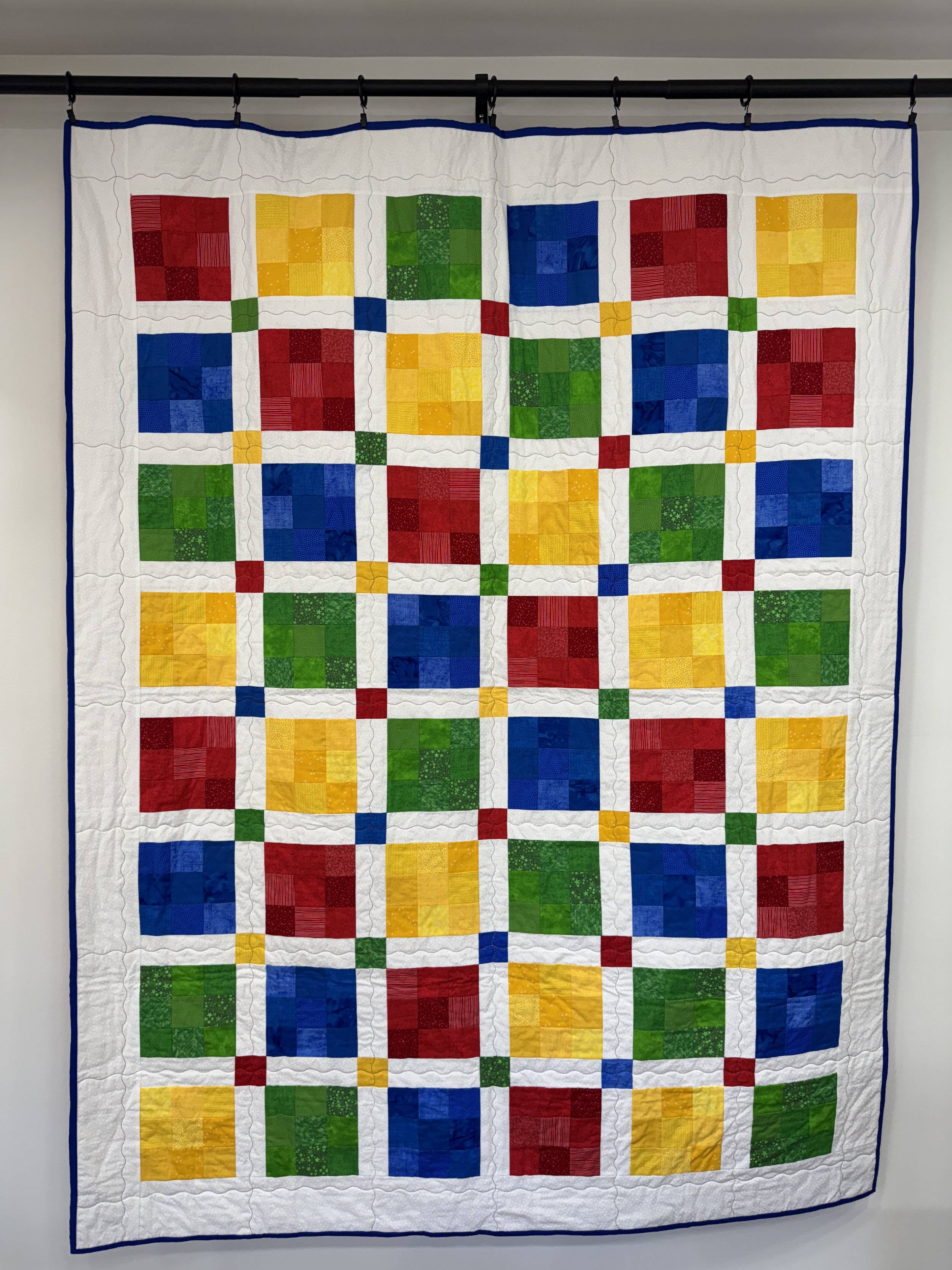 Colorful quilt with squares of red, yellow, green, and blue, bordered by white with a blue edge, hanging on a wall.