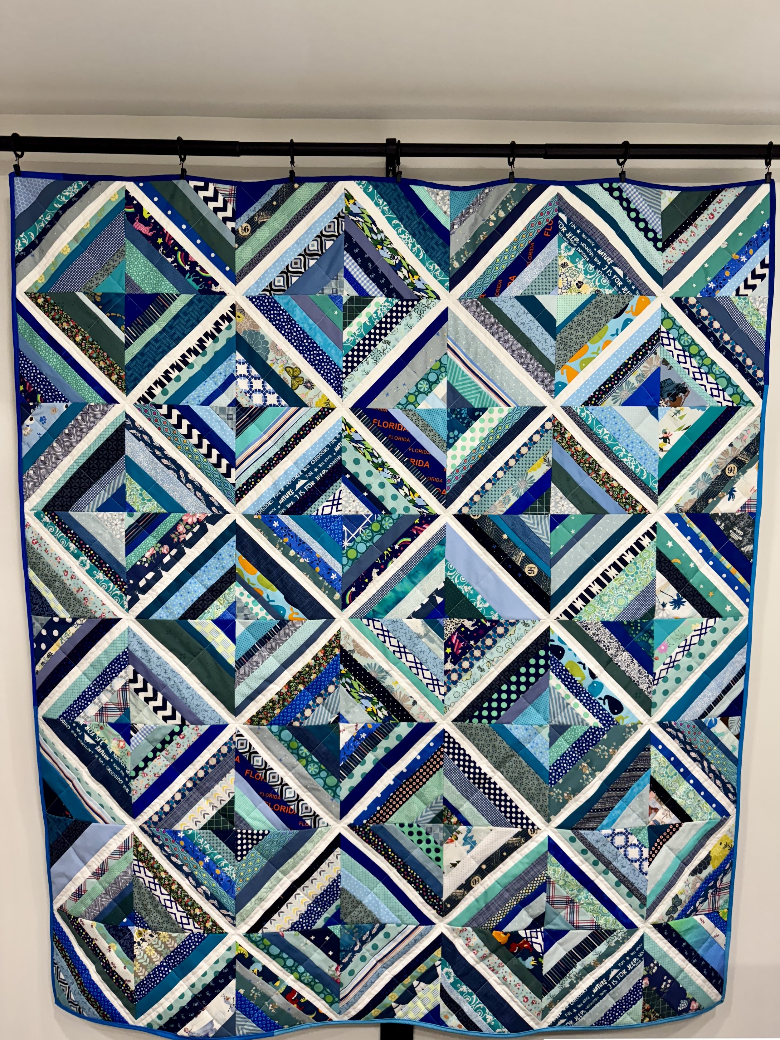 A string quilt made of numerous small fabric strips and triangles in shades of blue, green, and white, hanging on a black rod.