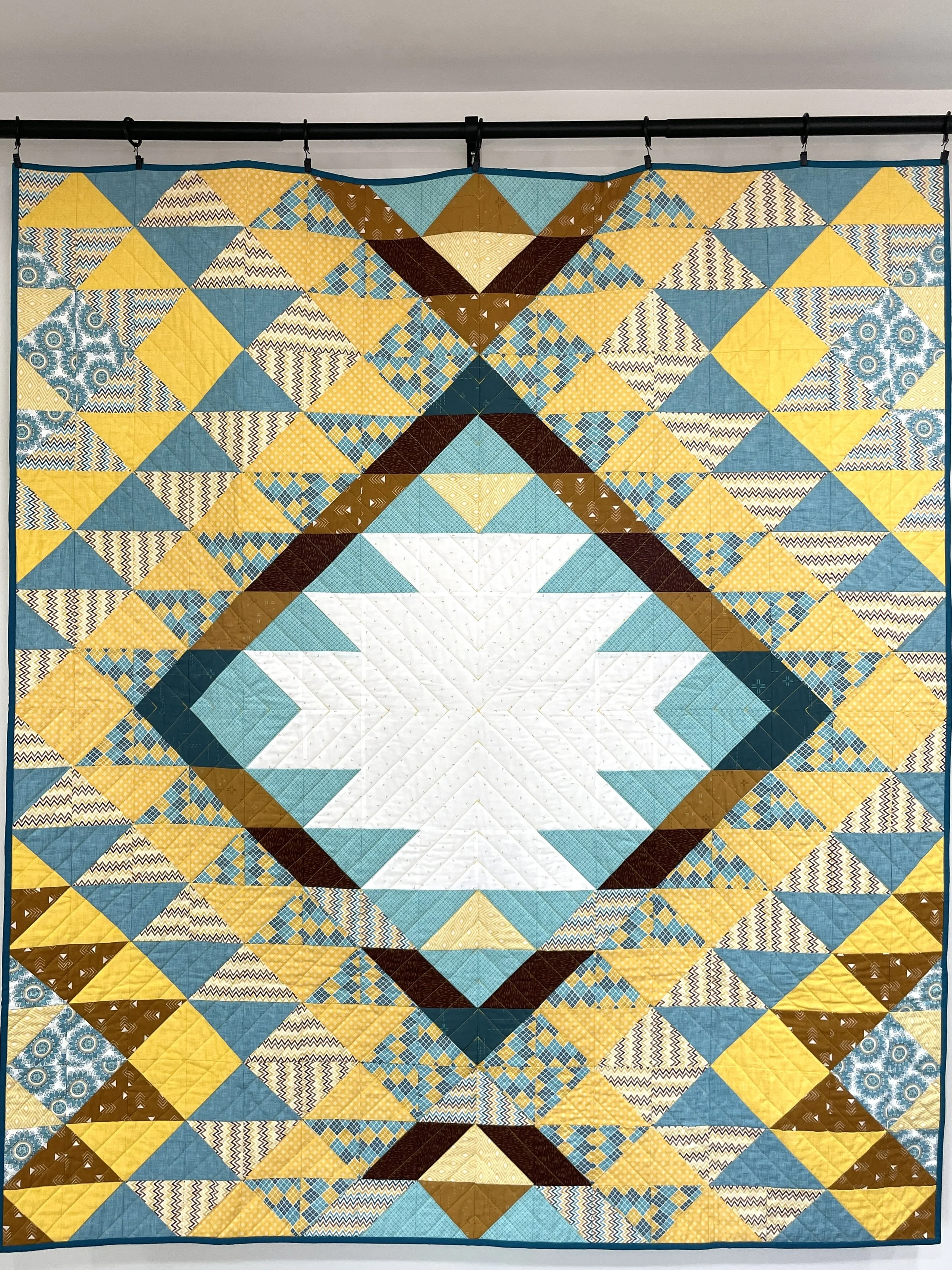 A colorful quilt with a geometric pattern consisting of triangles and diamond shapes in yellow, blue, white, and brown, displayed hanging on a black rod.