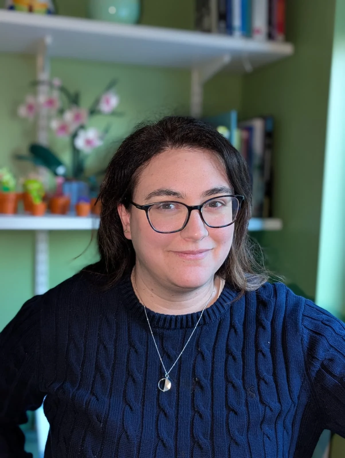 A woman with dark hair, glasses, and a navy blue sweater, smiling and sitting in a room with green walls, bookshelves, and potted plants in the background.