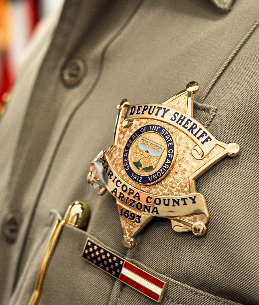 Close-up of a deputy sheriff badge on a gray uniform with an American flag pin on the pocket