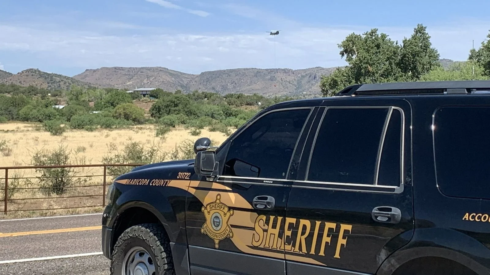 A Maricopa County sheriff's patrol vehicle parked on the side of a road with a desert landscape and mountains in the background. A helicopter is flying in the sky.