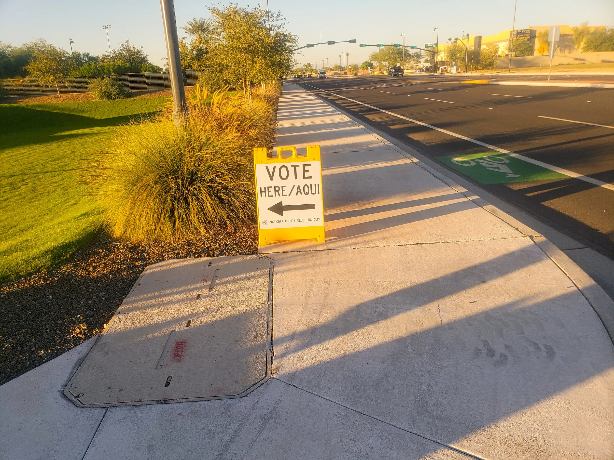 A sidewalk with a yellow sign pointing to the left that reads 'VOTE HERE/AQUI' and an arrow. The sidewalk is next to a crosswalk, a bike lane, and a grassy area with bushes and trees. The scene is lit by sunlight, with shadows cast on the sidewalk.