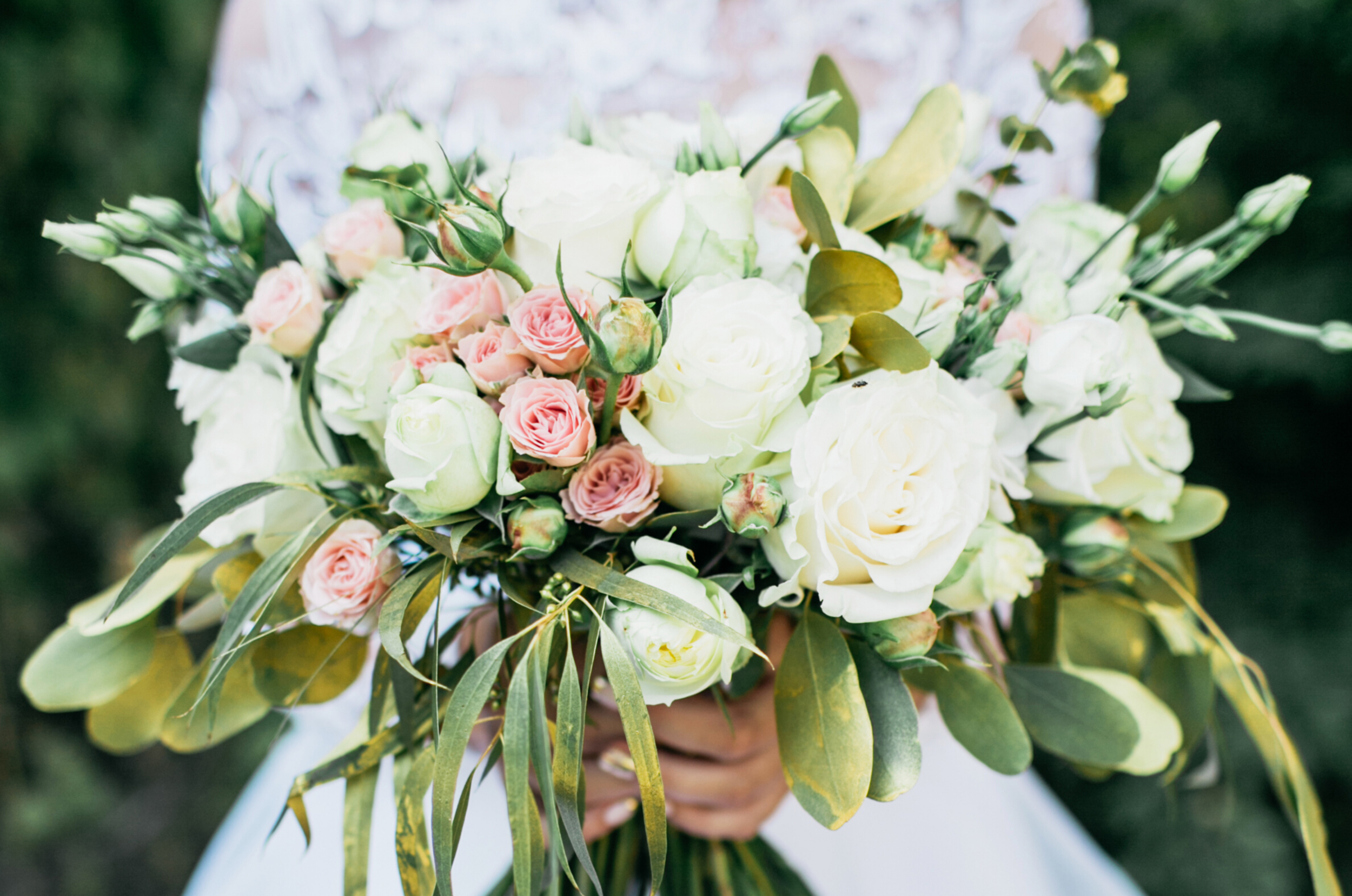 A bridal bouquet with white and pale pink roses, surrounded by green foliage, held by a woman in a white dress.