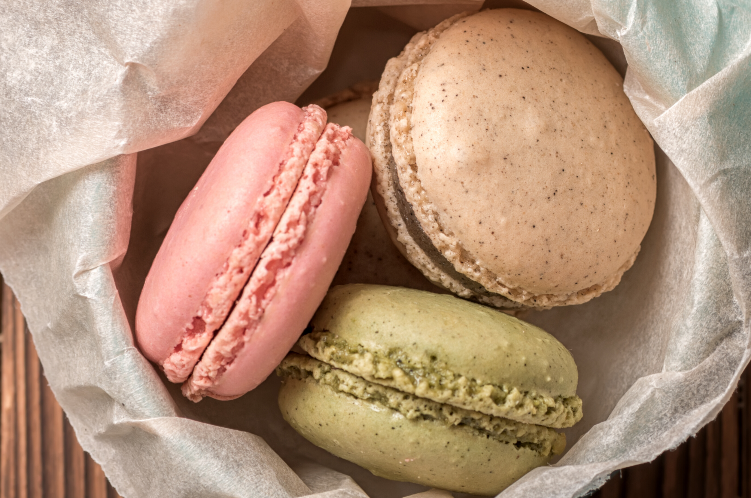 Three colorful macarons in a basket lined with parchment paper, placed on a wooden surface.