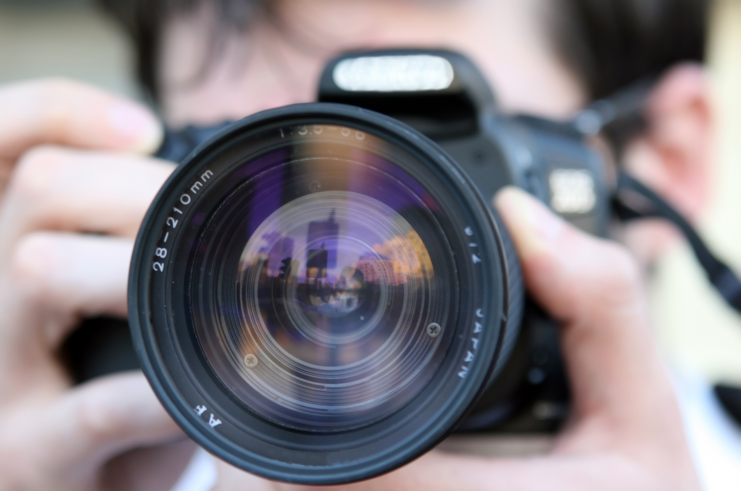 A person holding a camera with the lens pointed directly at the viewer, showing a reflection of skyline and sky in the lens.
