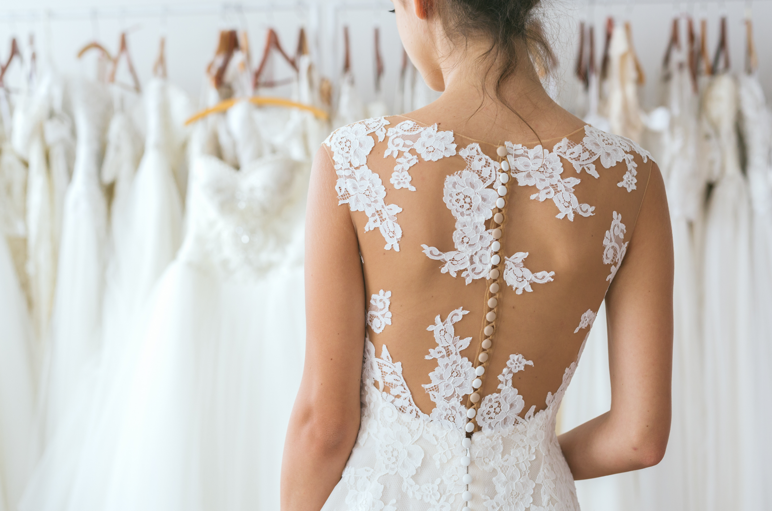 A woman trying on a wedding dress with a sheer lace back and buttons, surrounded by other wedding dresses on display.