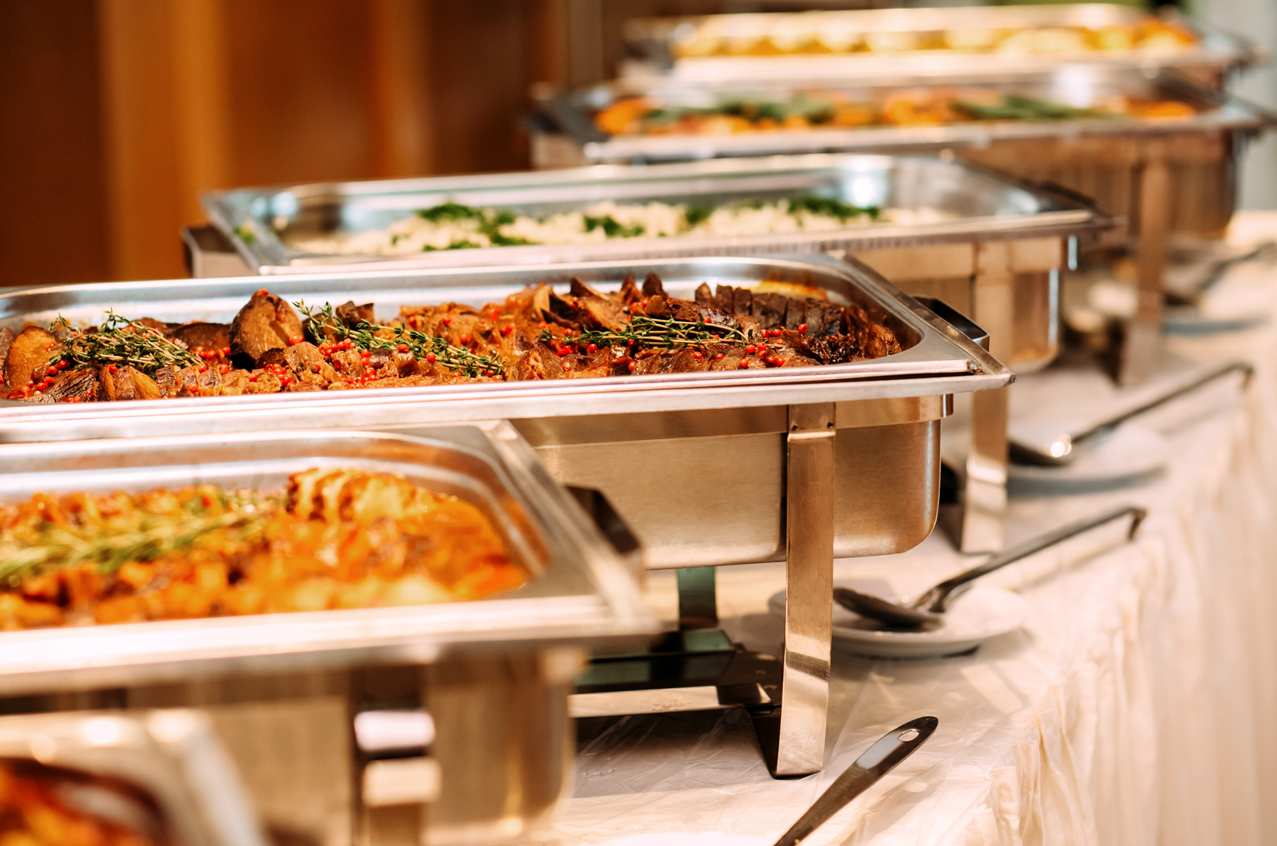 Chafing dishes filled with various hot foods, including roasted meats and vegetables, arranged on a buffet table with serving utensils.