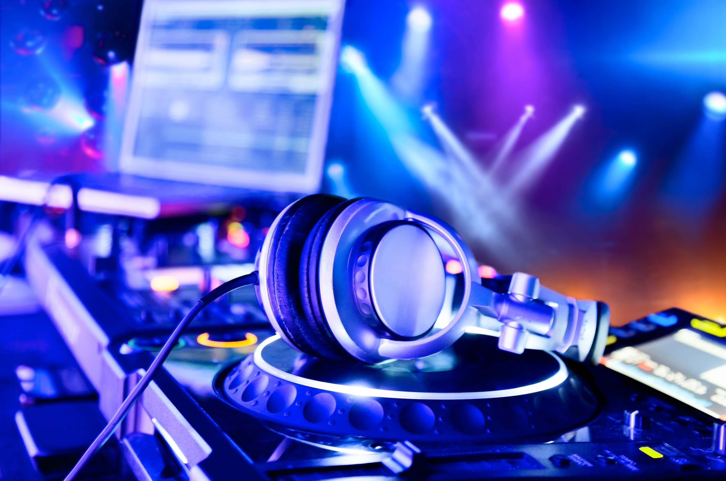DJ headphones resting on a DJ turntable with a glowing ring, with colorful stage lights and DJ equipment in the background.
