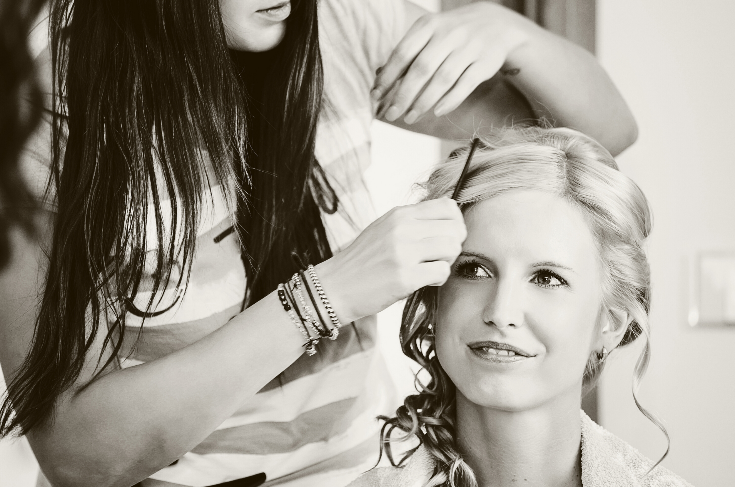 A woman with light-colored hair getting her hair styled by a stylist with dark hair, who is making final touches on her hair.
