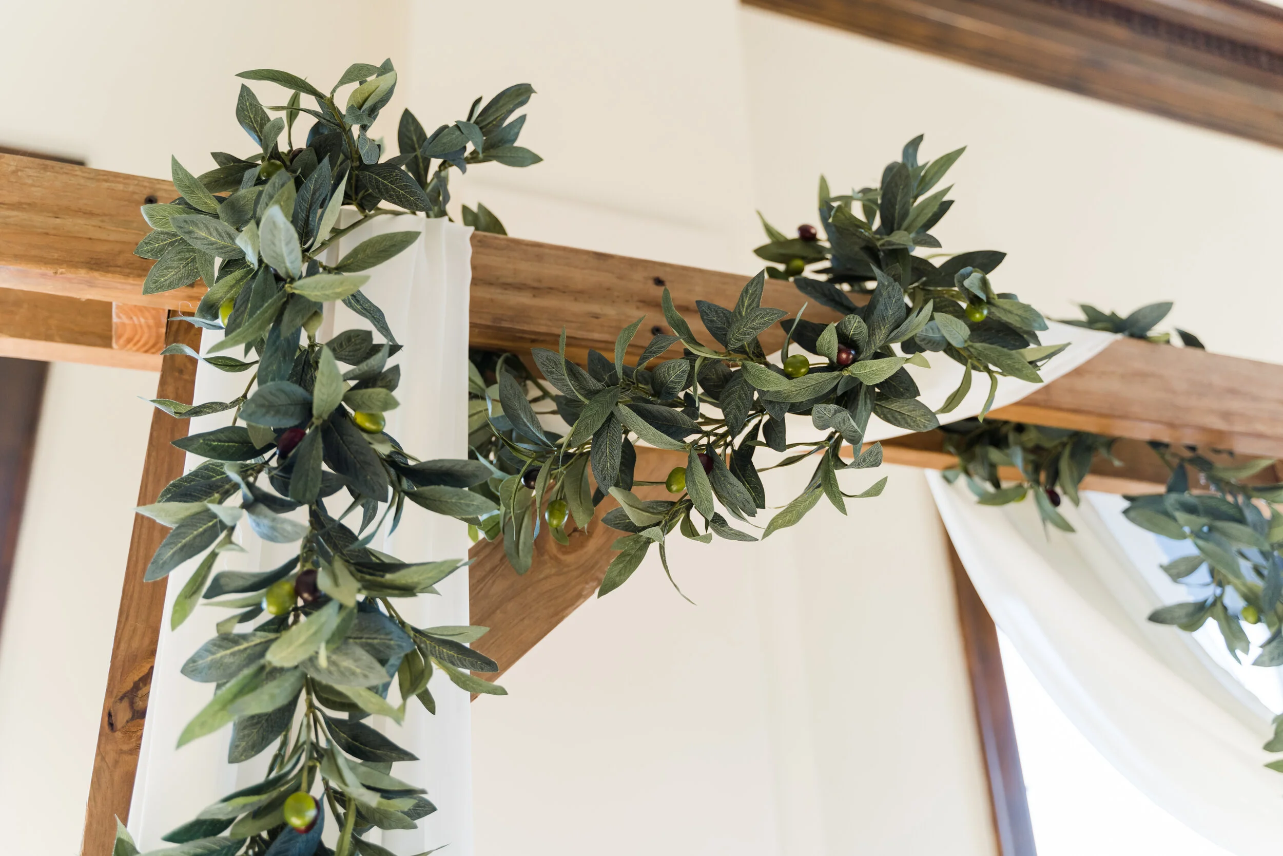 Decorative green leafy garland draped along a wooden beam with white fabric accents in a room.
