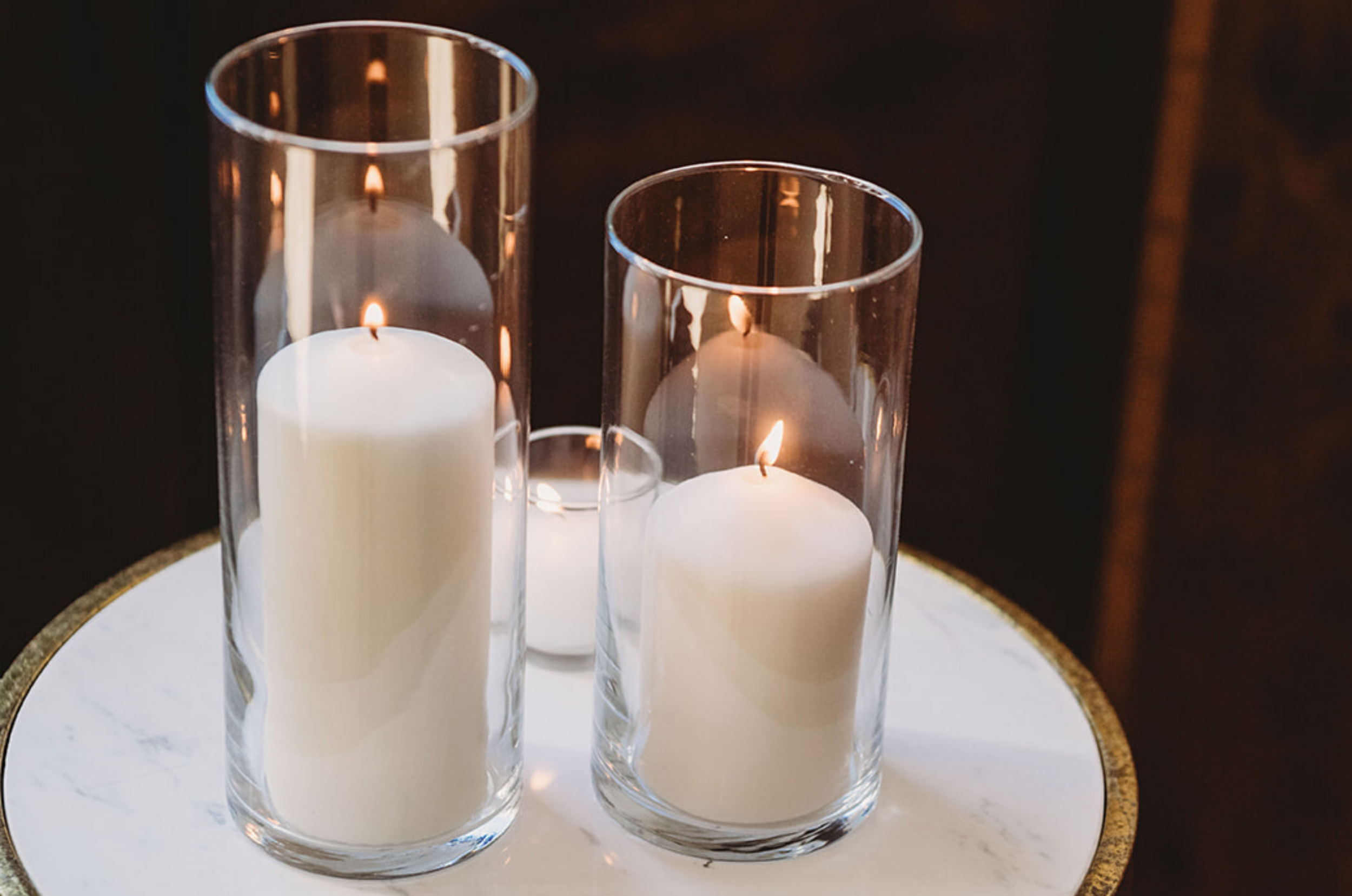 Three glass candle holders with white pillar candles lit inside, placed on a white round table with a gold rim.